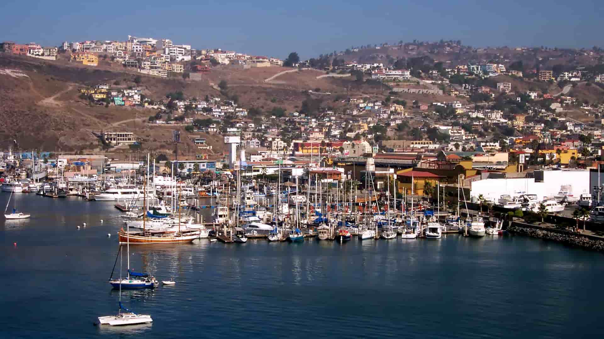 A bustling marina in Ensenada, Mexico, filled with sailboats and yachts, with the colorful buildings of the city climbing up a barren hillside in the background.