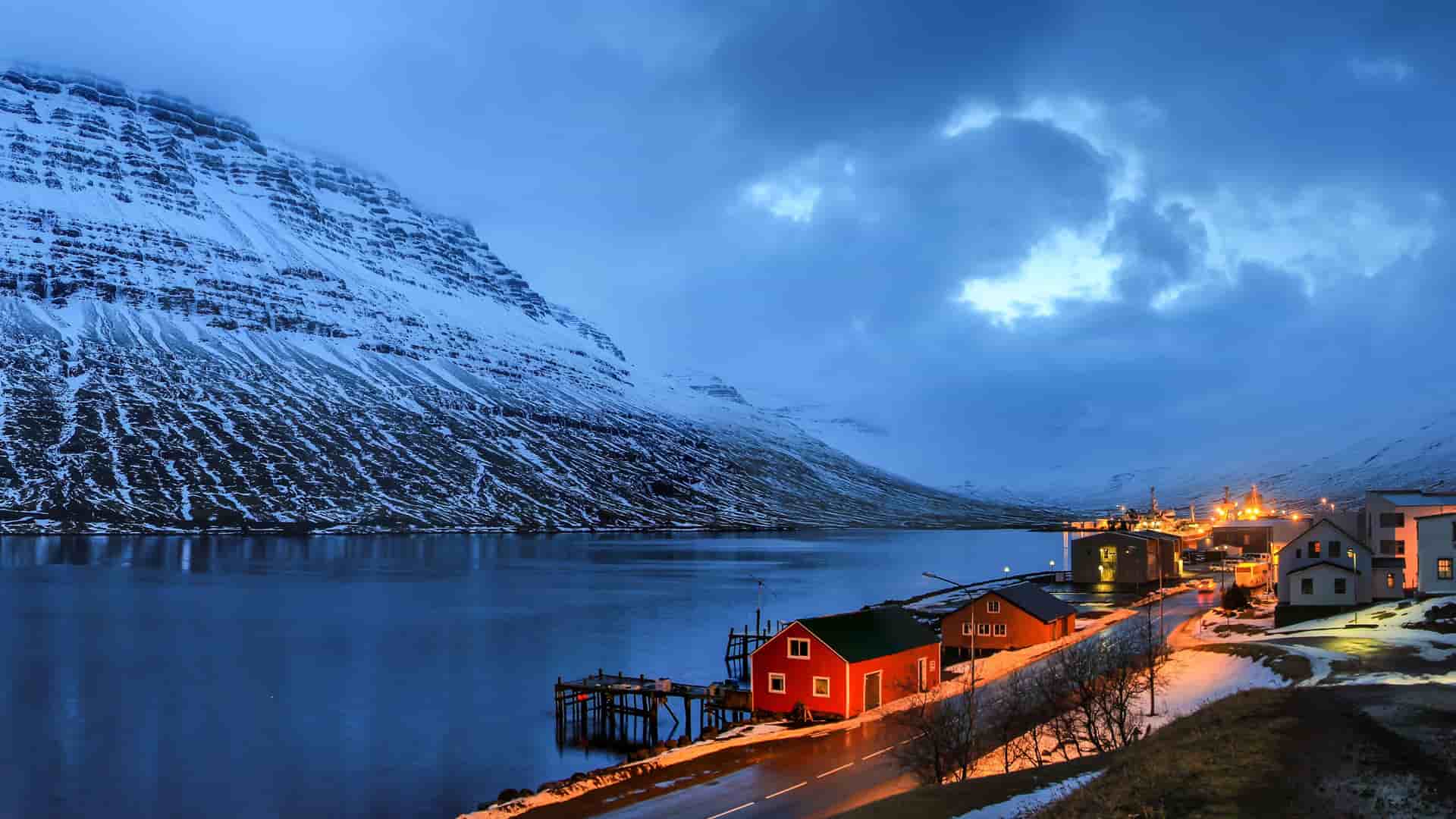A dramatic twilight shot of Eskifjordur, Iceland, with a small cluster of illuminated buildings nestled on the shore of a calm fjord, surrounded by a towering, snow-covered mountain.
