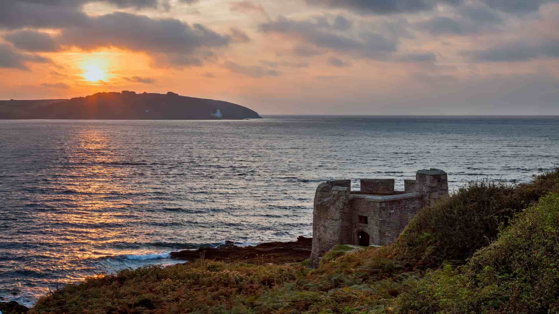 A breathtaking sunset over the sea near Falmouth, Cornwall, with a historical stone fort in the foreground and a distant coastline silhouetted against the bright, cloudy sky.