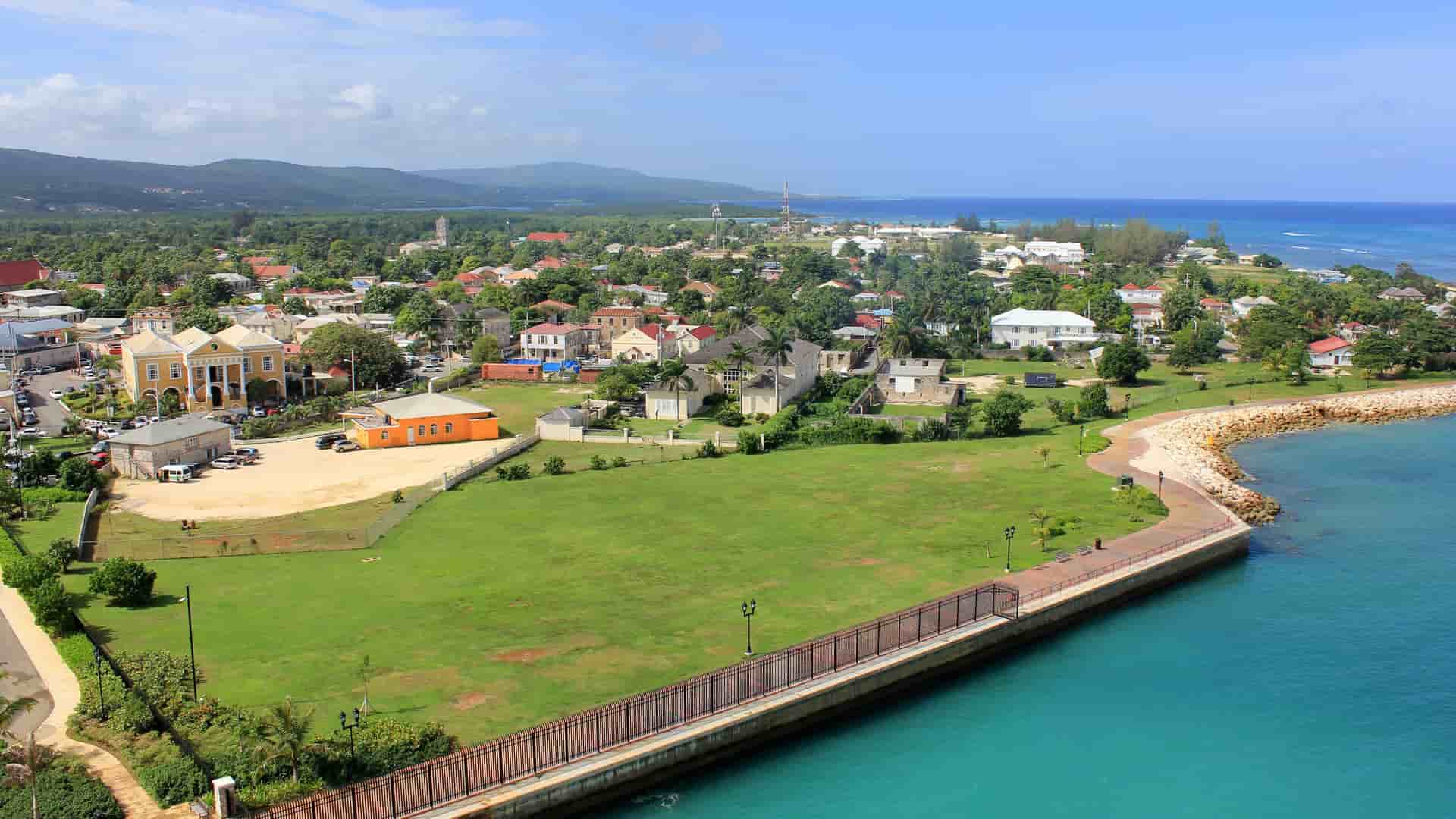 An aerial view of the historic town of Falmouth, Jamaica, showing a coastal promenade along the turquoise sea, colorful colonial-era buildings, and lush green hills in the background.