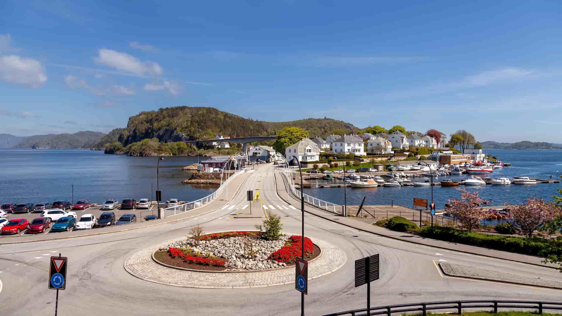 "A sunny panoramic shot of Farsund, Norway, with a busy roundabout in the foreground leading to a bridge connecting the mainland to a small island filled with white houses, a bustling marina, and lush green hills.  "