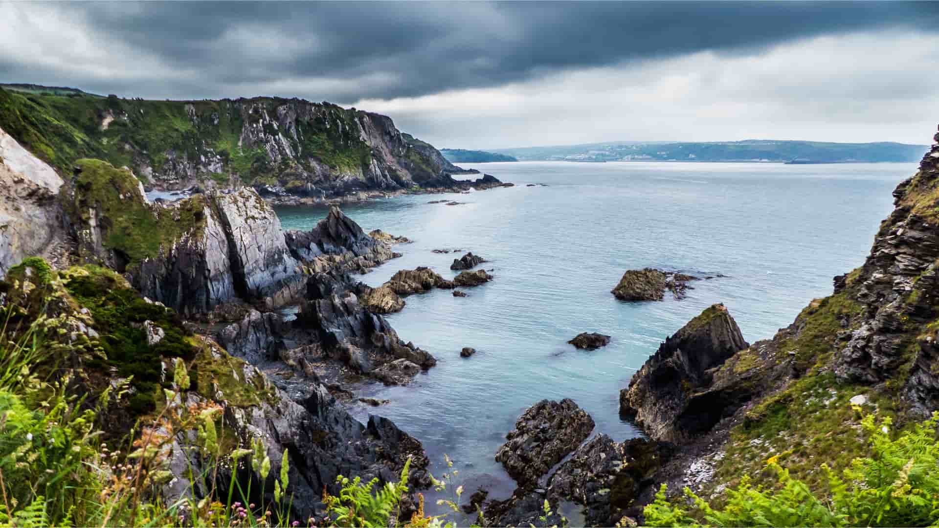 A moody and dramatic landscape of Fishguard, Wales, featuring a rugged coastline with sharp, rocky cliffs and a serene blue bay under a heavy, cloudy sky.