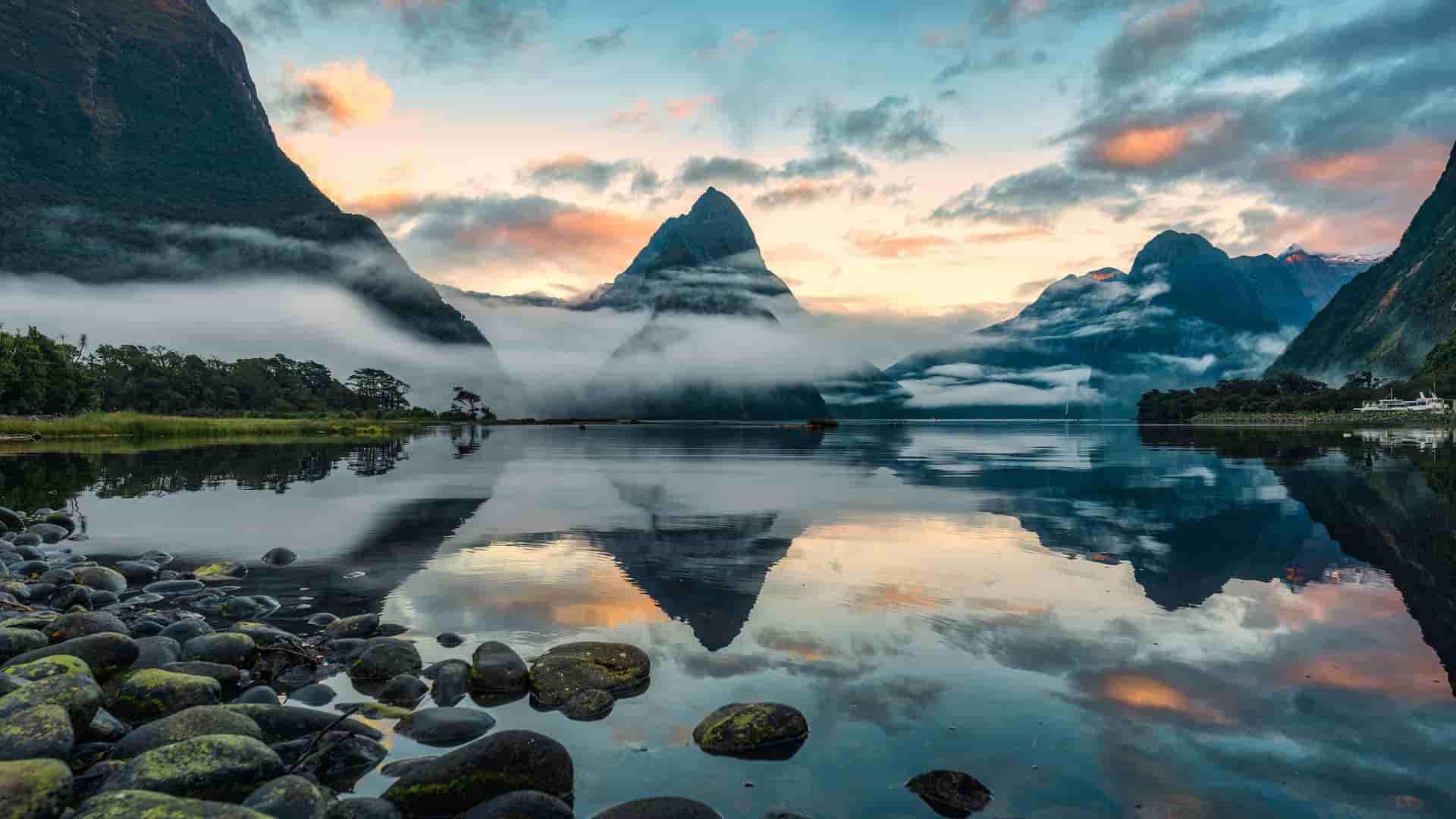A serene sunrise view of Milford Sound in Fiordland National Park, New Zealand, with the iconic Mitre Peak reflected in the perfectly still water and clouds hugging the surrounding mountains.