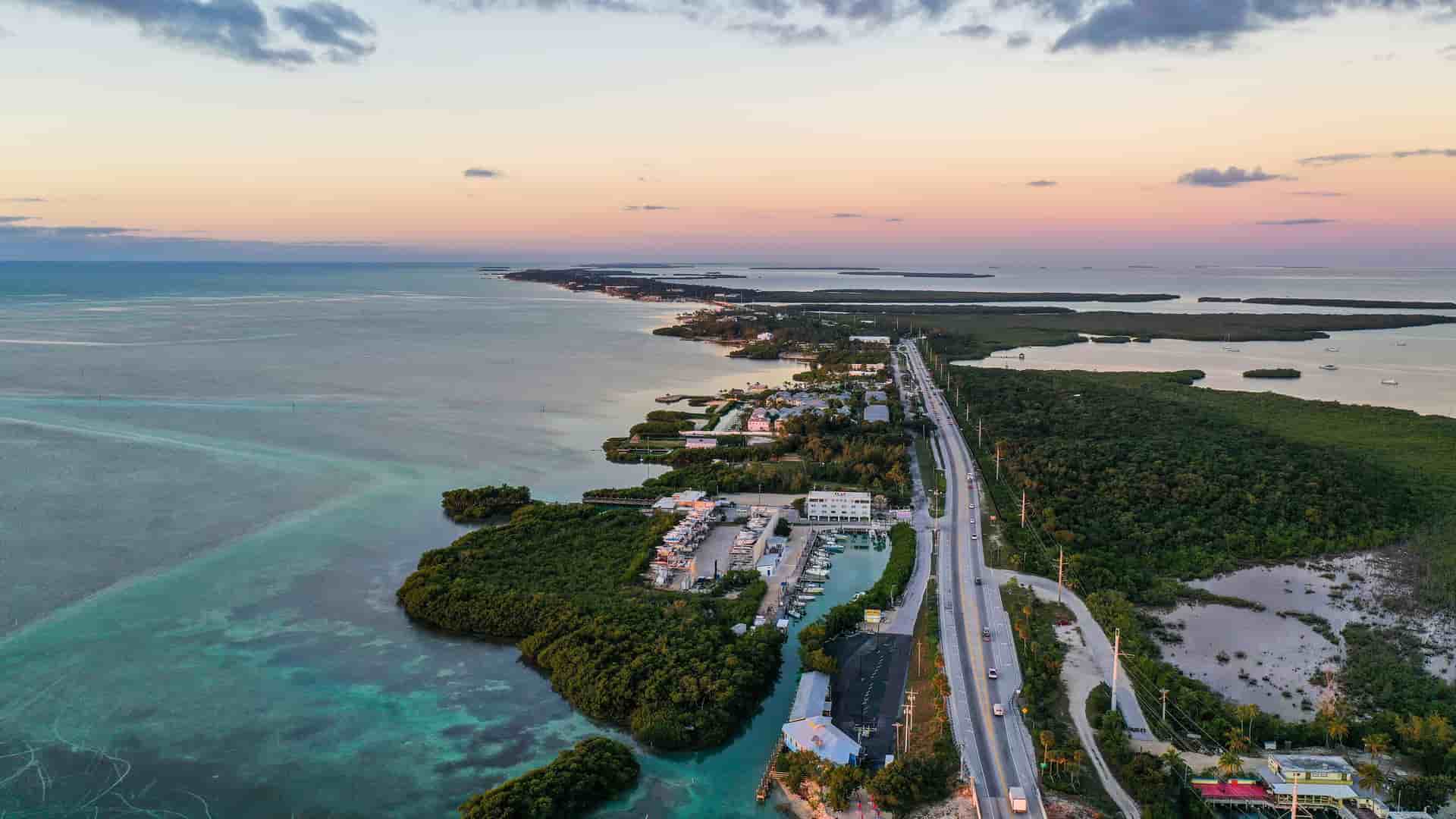 An aerial shot of the Florida Keys at sunset, showing a long highway stretching along the thin strip of land, with the calm, turquoise Florida Strait on one side and a lush mangrove coastline on the other.