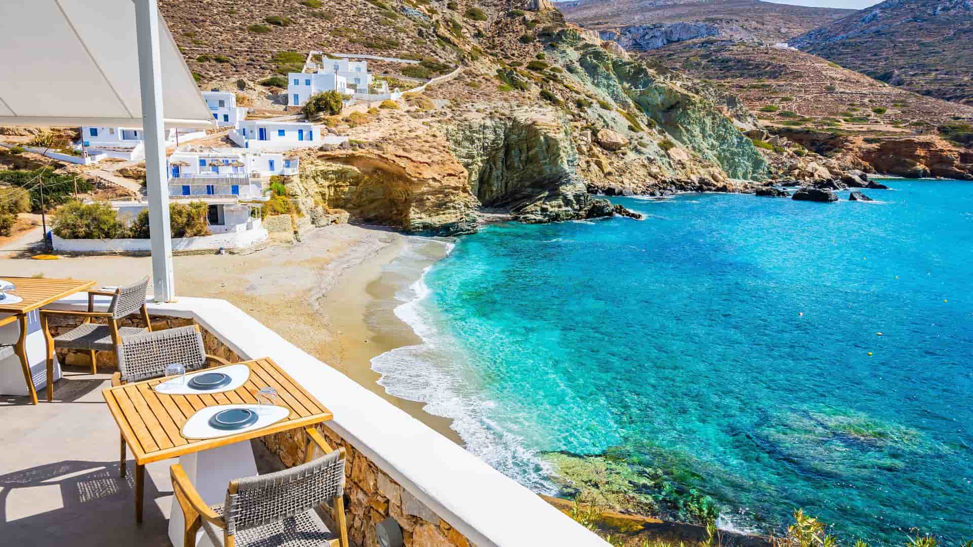 A high-angle view from a restaurant patio overlooking the stunning turquoise waters of a secluded bay and a small sandy beach on the picturesque island of Folegandros in Greece.