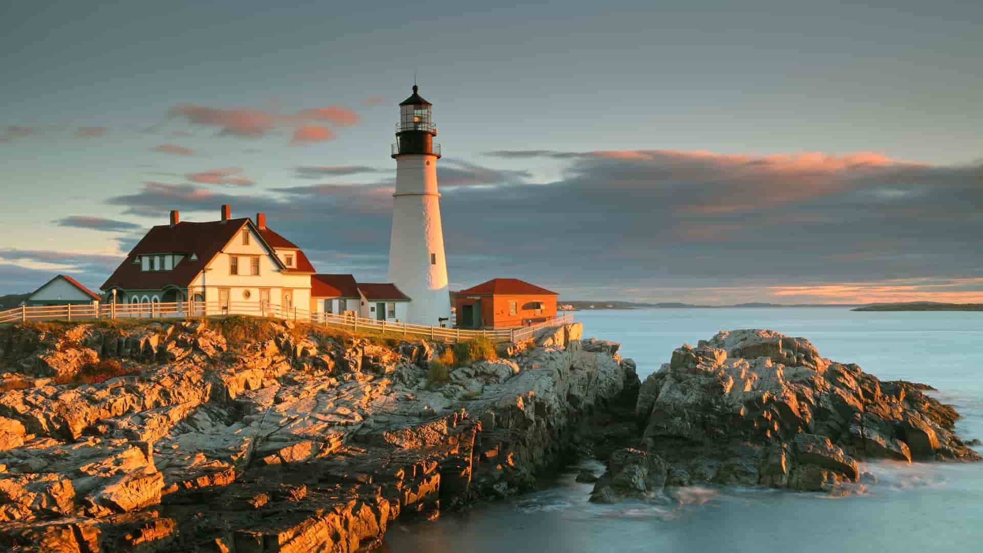 A picturesque landscape shot of the iconic Portland Head Light lighthouse at sunset, with a beautiful golden glow on the rocky coastline and a calm ocean.