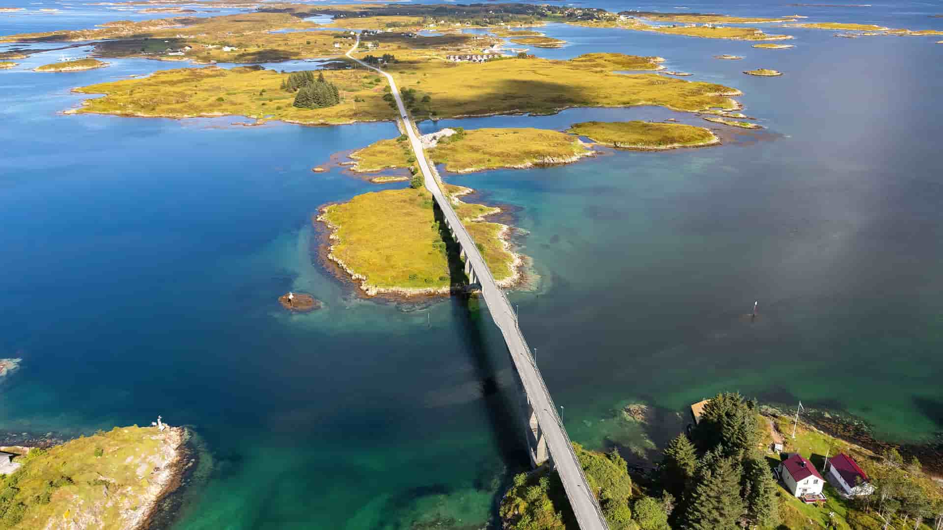 An aerial view of a winding road connecting small, grassy islands in the Froya region of Norway, surrounded by the clear blue and green waters of the sea.