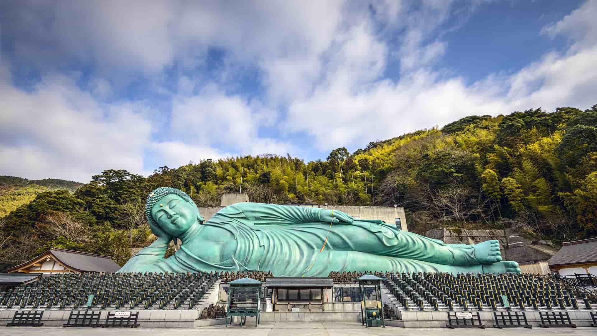 A panoramic shot of the enormous, reclining bronze Buddha statue at Nanzoin Temple in Fukuoka, Japan, with green trees and a building in the background.