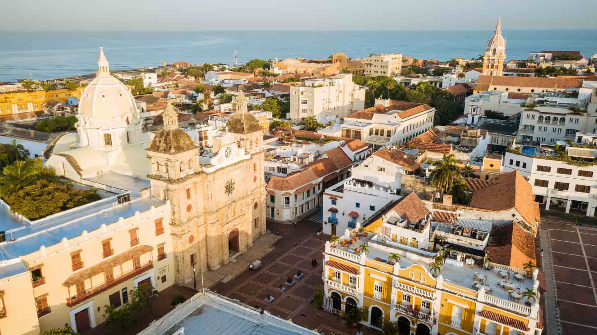 An aerial view of the historic walled city of Cartagena, Colombia, showing the vibrant architecture of churches and colonial buildings, with the ocean in the background under a soft morning light.