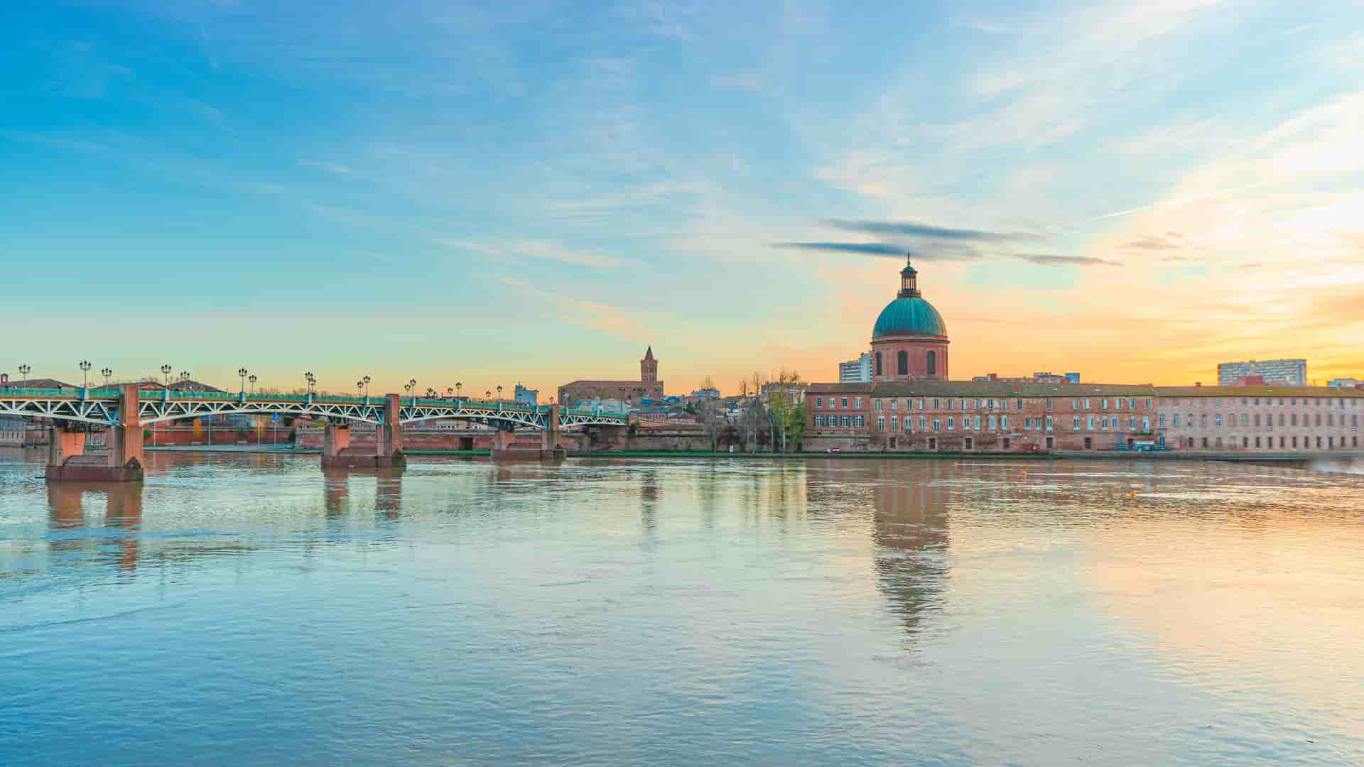 A beautiful twilight shot of the Garonne River in Toulouse, France, featuring a bridge and the dome of the Hôpital de La Grave in the background.