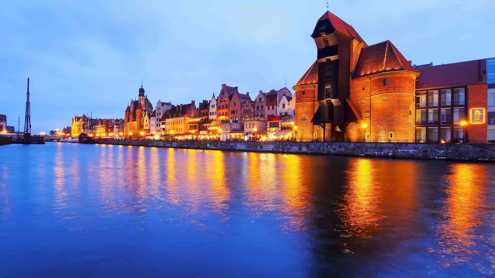 A scenic twilight view of Gdansk, Poland, showcasing the historic waterfront with its colorful buildings and the iconic medieval crane, beautifully reflected in the Motlawa River.