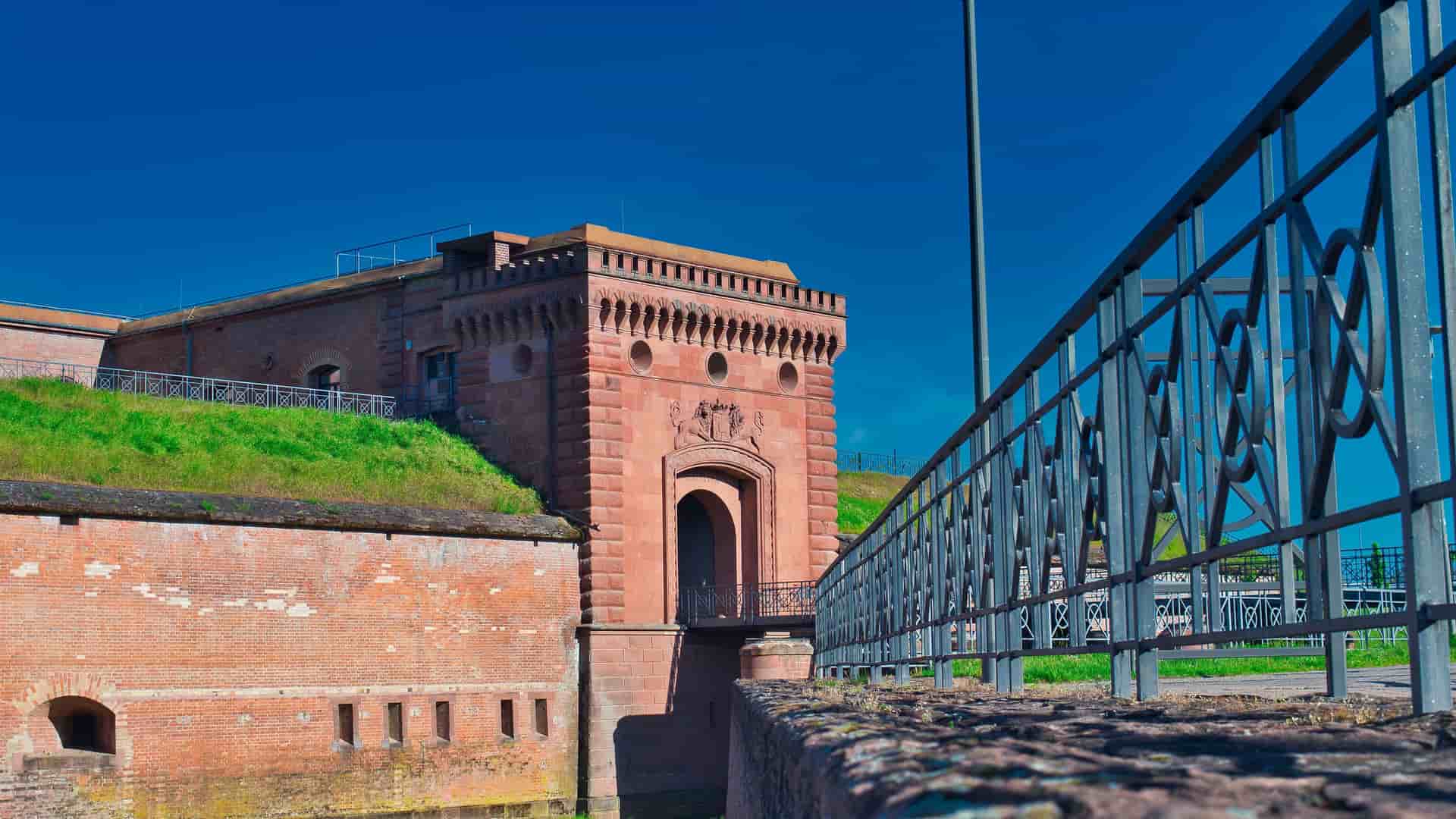 A beautiful wide-angle shot of the historic Festung Germersheim fortress in Germany, highlighting the red brick building with its arched entrance, a moat, and green grass on top.