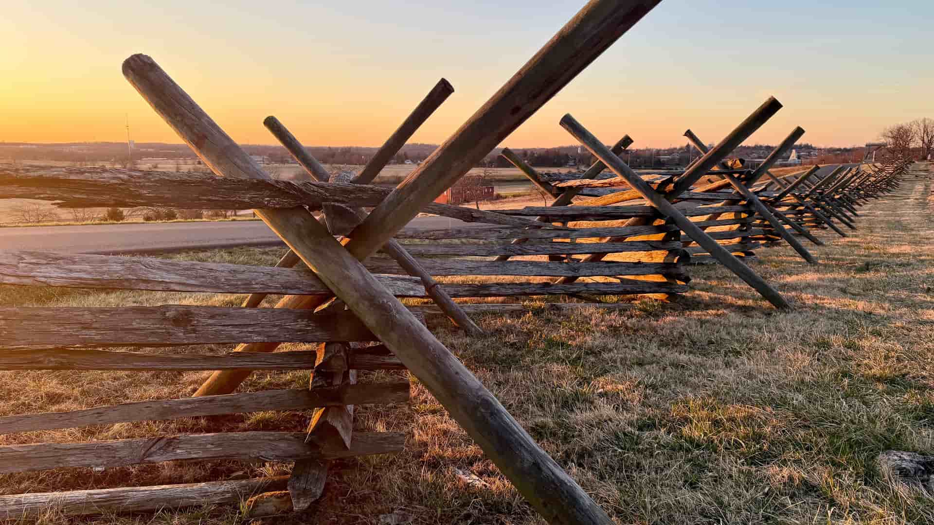 A scenic ground-level shot of a historic wooden split-rail fence on a grassy field at the Gettysburg National Military Park at sunset, with golden light illuminating the landscape.