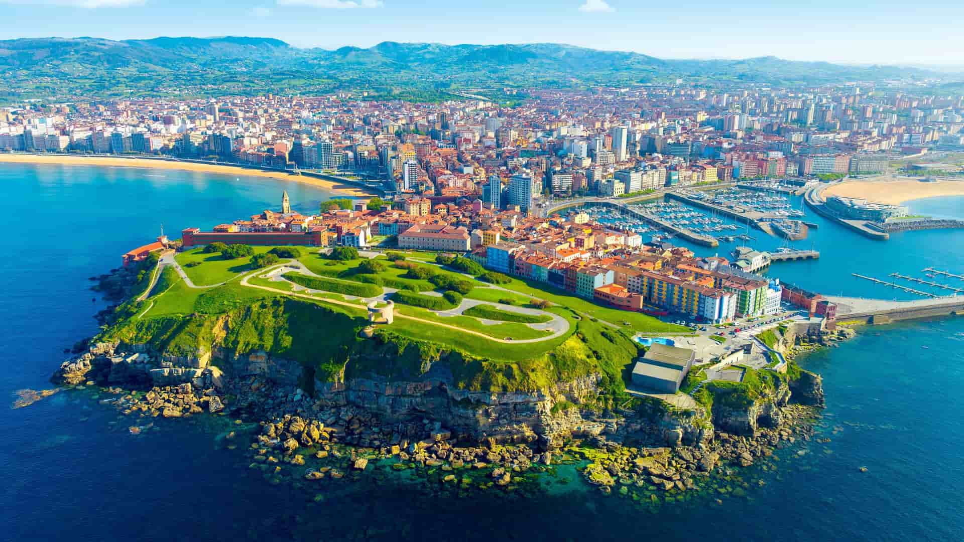 An aerial shot of Gijon, Spain, showcasing the Cimavilla Peninsula with green grass and cliffs, and the beautiful city and marina in the background.