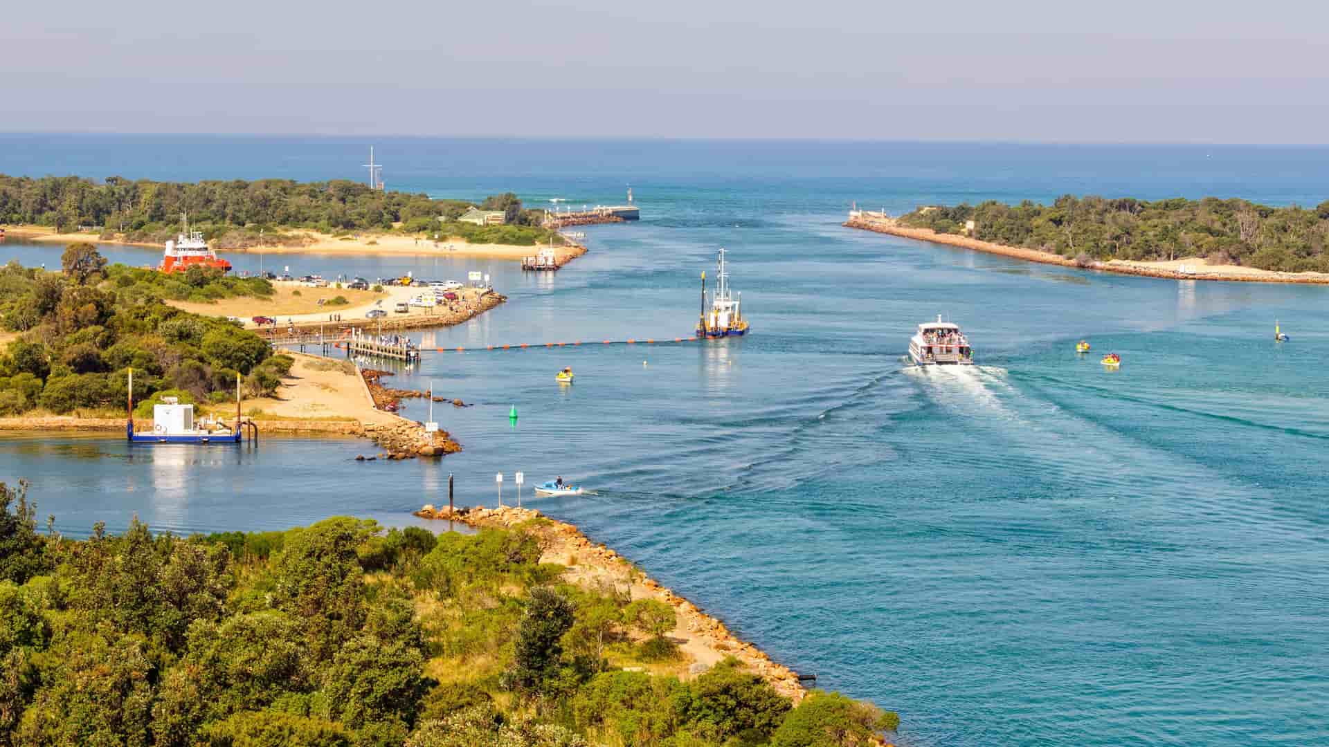 A beautiful wide-angle shot of the Gippsland Coast in Victoria, Australia, showcasing the meeting point of the lake and the ocean with boats sailing in the calm water.