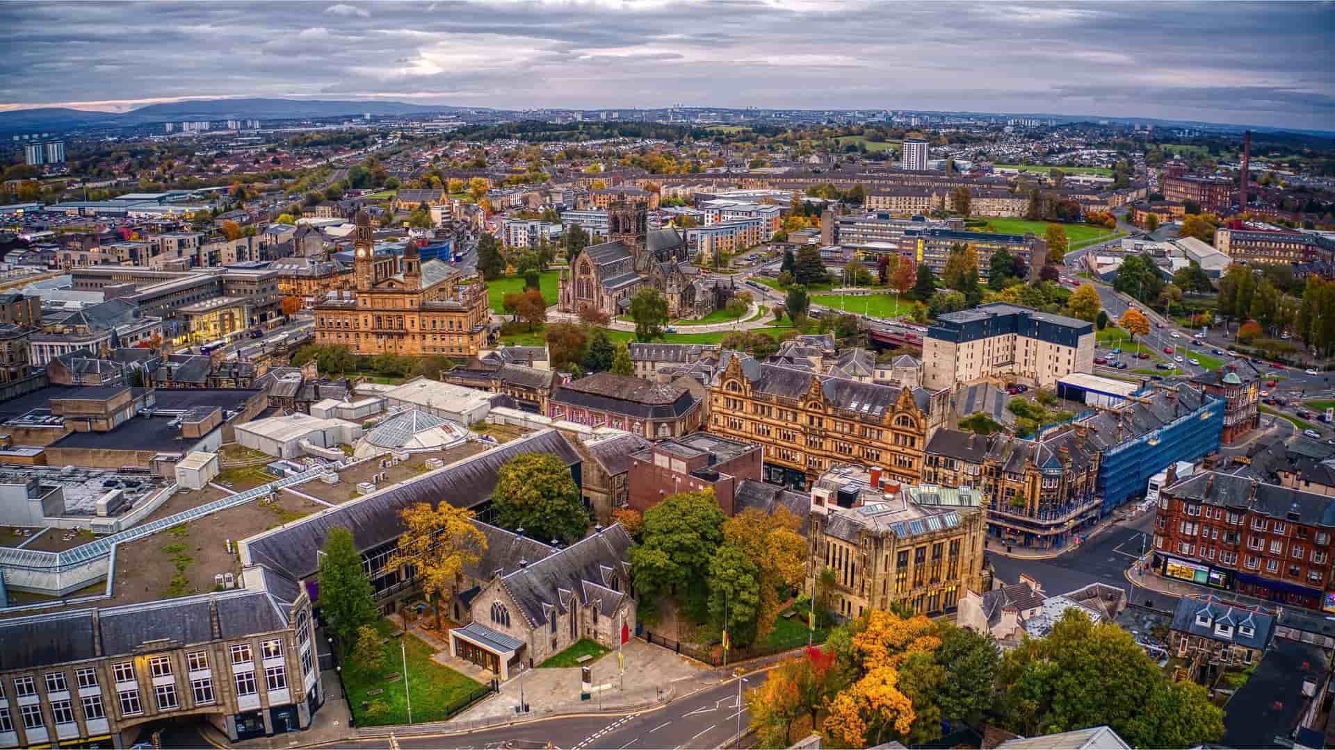 An aerial shot of Glasgow, Scotland, featuring the city's historic architecture, including the University of Glasgow, and a large urban landscape filled with autumn-colored trees under a dramatic, cloudy sky.