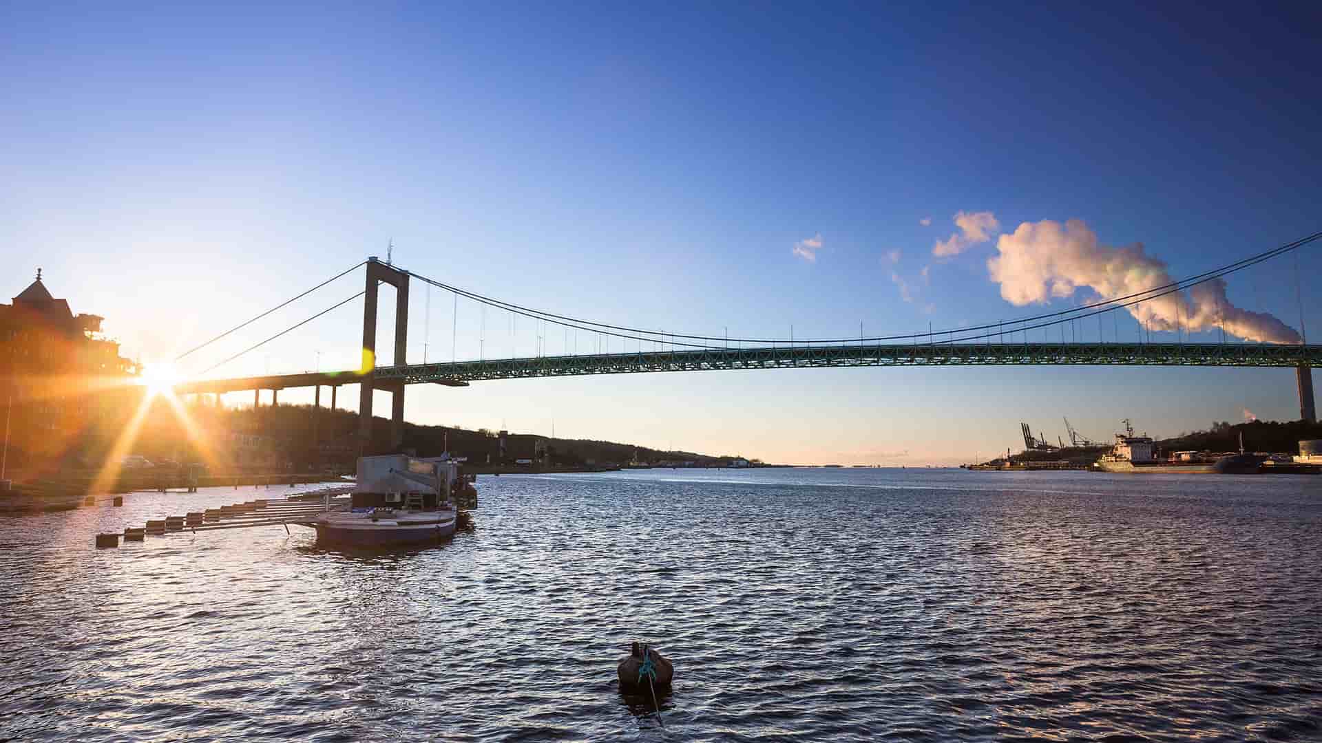 A wide-angle sunset view of the Älvsborg Bridge in Gothenburg, Sweden, spanning over the Göta Älv river with ships and a port visible in the background.