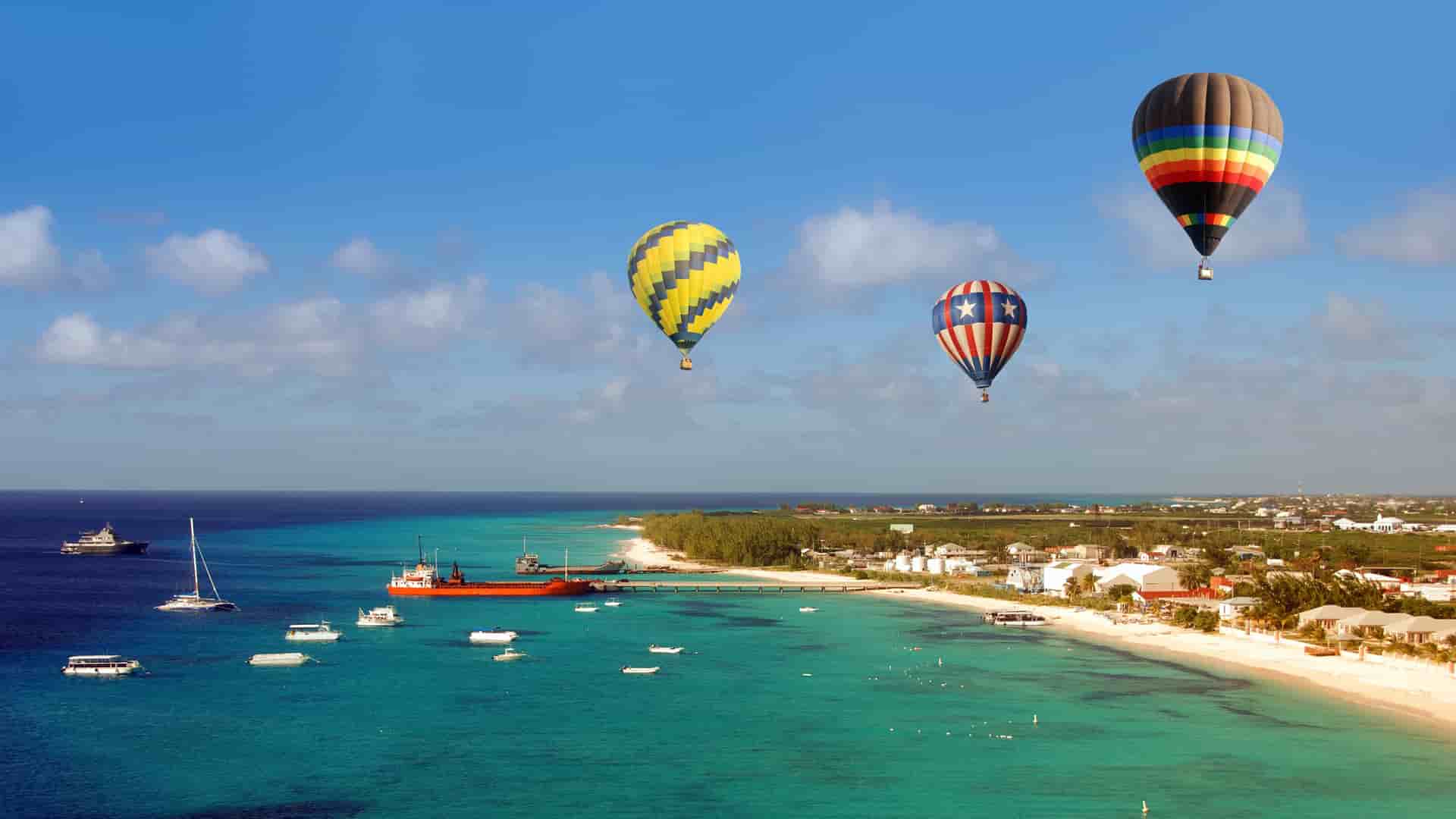 A sunny, tropical beach on Grand Turk Island with multiple hot air balloons flying over the turquoise water and shoreline.