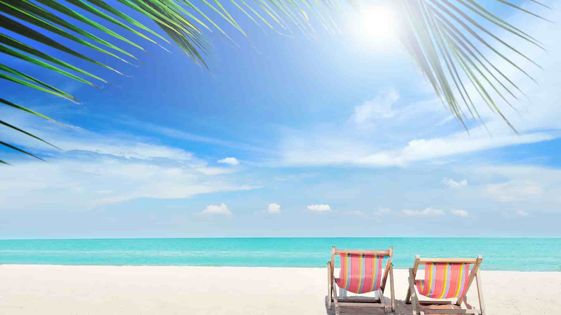 Two colorful striped beach chairs sit on the white sand beach of Great Stirrup Cay, Bahamas, facing the clear turquoise water under a bright blue sky with palm fronds overhead.