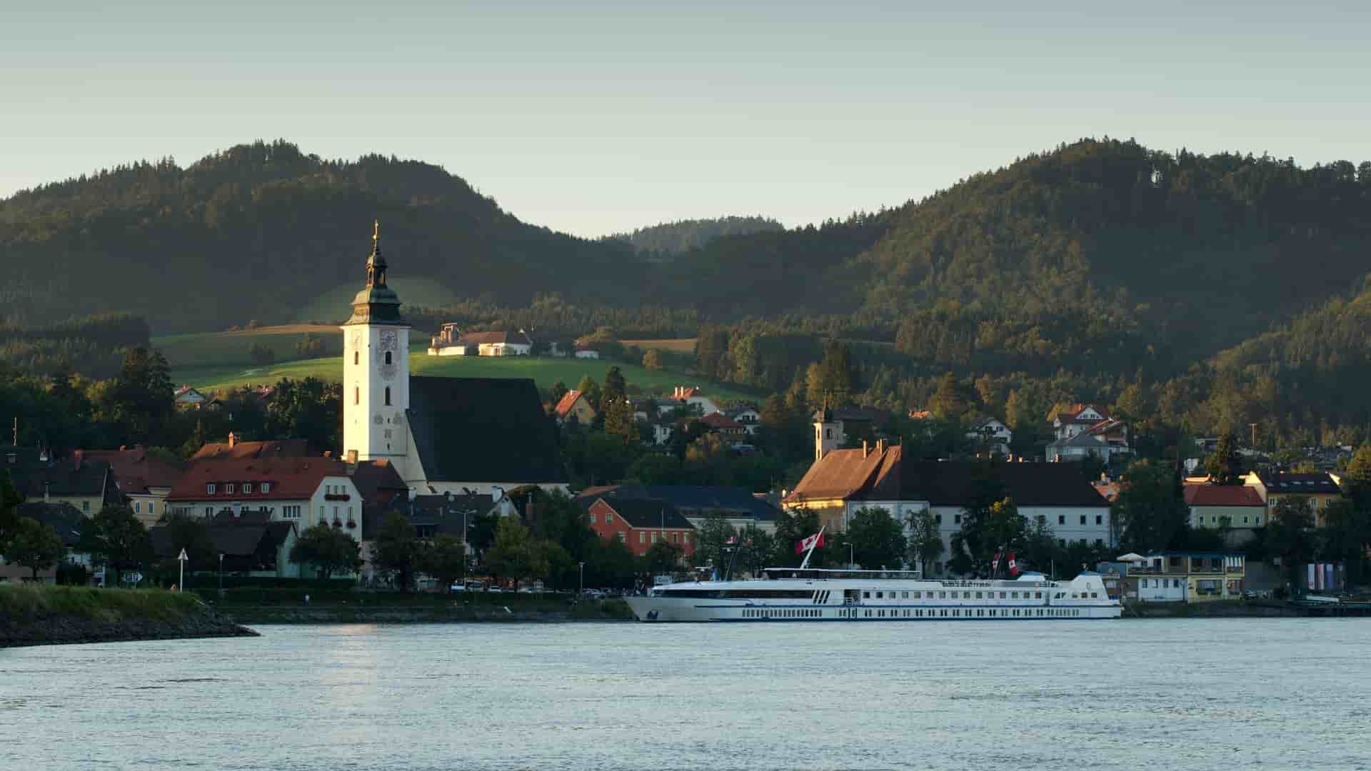 A river cruise ship docked in the picturesque town of Grein, Austria, with a prominent church steeple and residential buildings nestled against lush green hills.