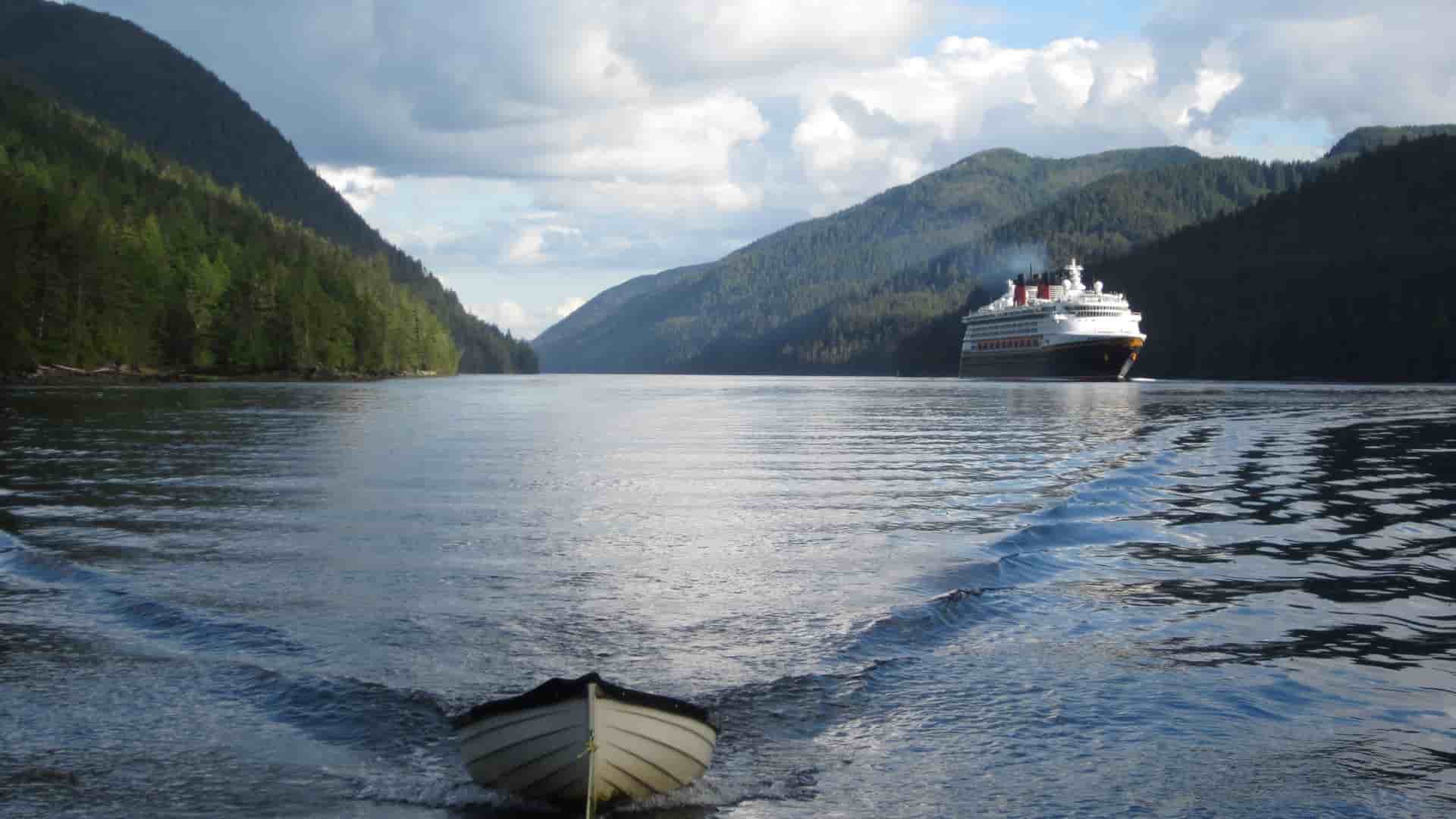 A large cruise ship sailing through the scenic Grenville Channel in British Columbia, with a small boat in the foreground and mountains covered in lush green forests on both sides.