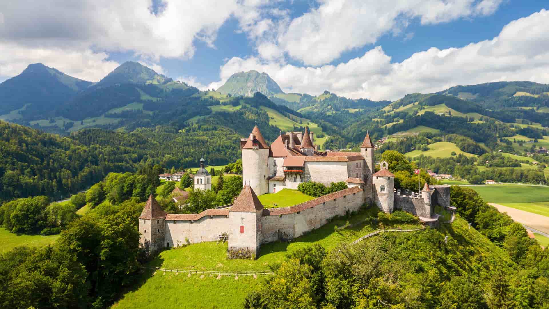 An aerial view of the historic Gruyères Castle, with its red-roofed towers, nestled on a lush green hillside in the scenic Swiss countryside, surrounded by mountains.