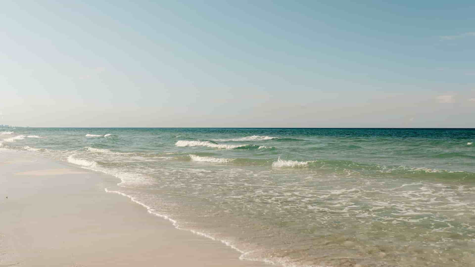 A tranquil wide-angle shot of a sandy beach along the Gulf of Mexico, with gentle waves rolling onto the shore and a clear blue sky.