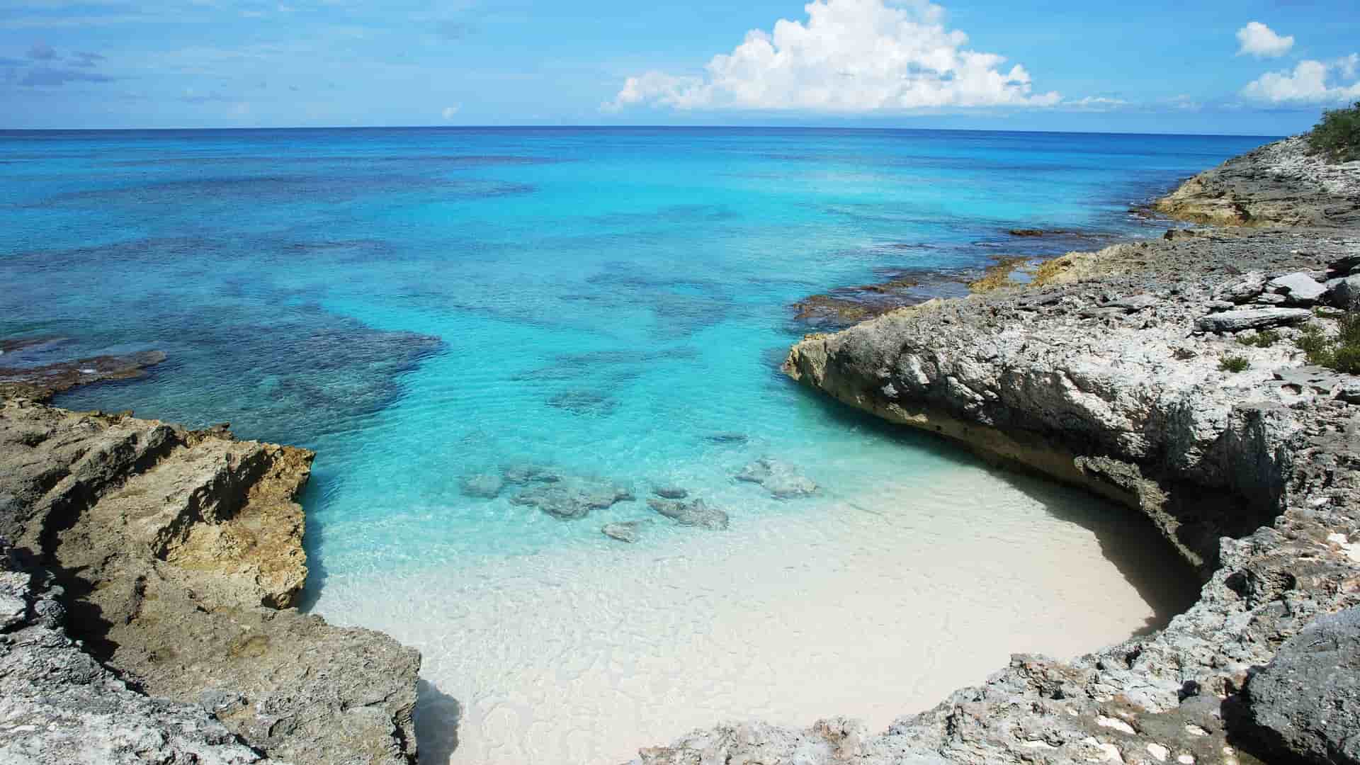 A secluded cove with white sand and turquoise water on Half Moon Cay, Bahamas, surrounded by jagged, rocky cliffs and a serene blue ocean under a partly cloudy sky.