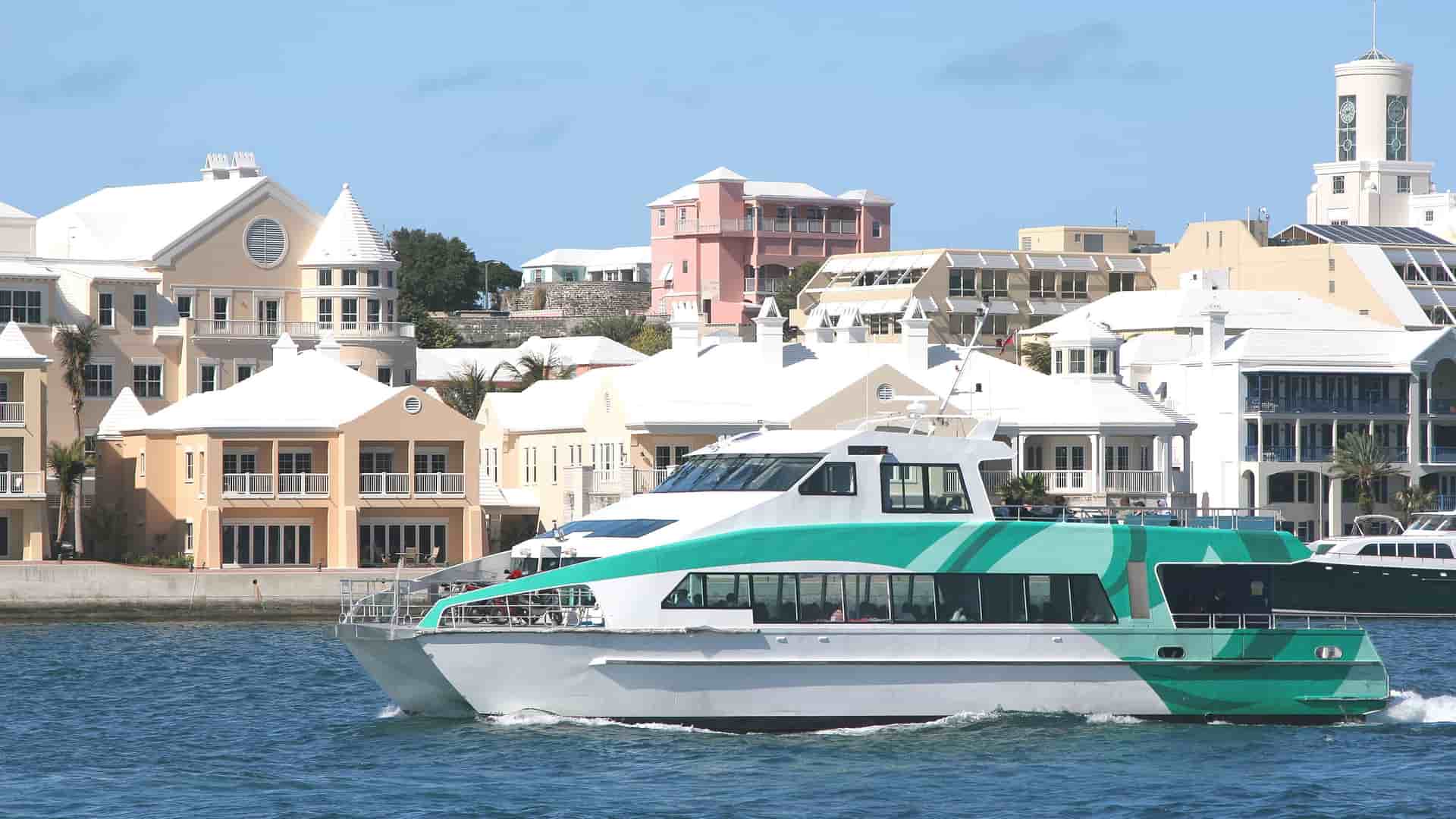 A turquoise and white ferry boat cruises through the harbor in Hamilton, Bermuda, with a backdrop of pastel-colored buildings and houses along the waterfront.