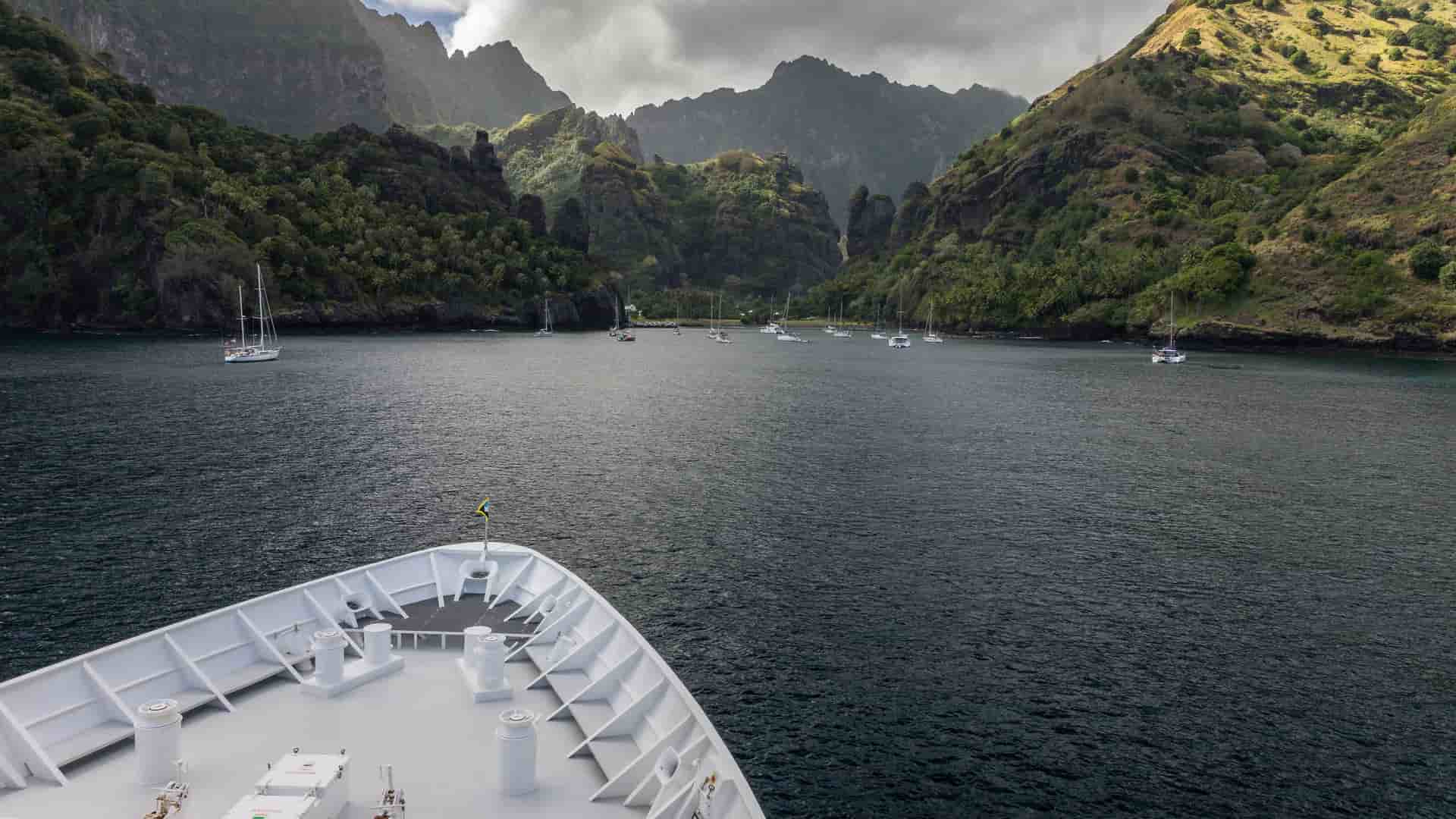 A dramatic view from the bow of a ship entering Hanavave Bay, Fatu Hiva, with numerous sailboats anchored in the dark water surrounded by lush, steep volcanic mountains.
