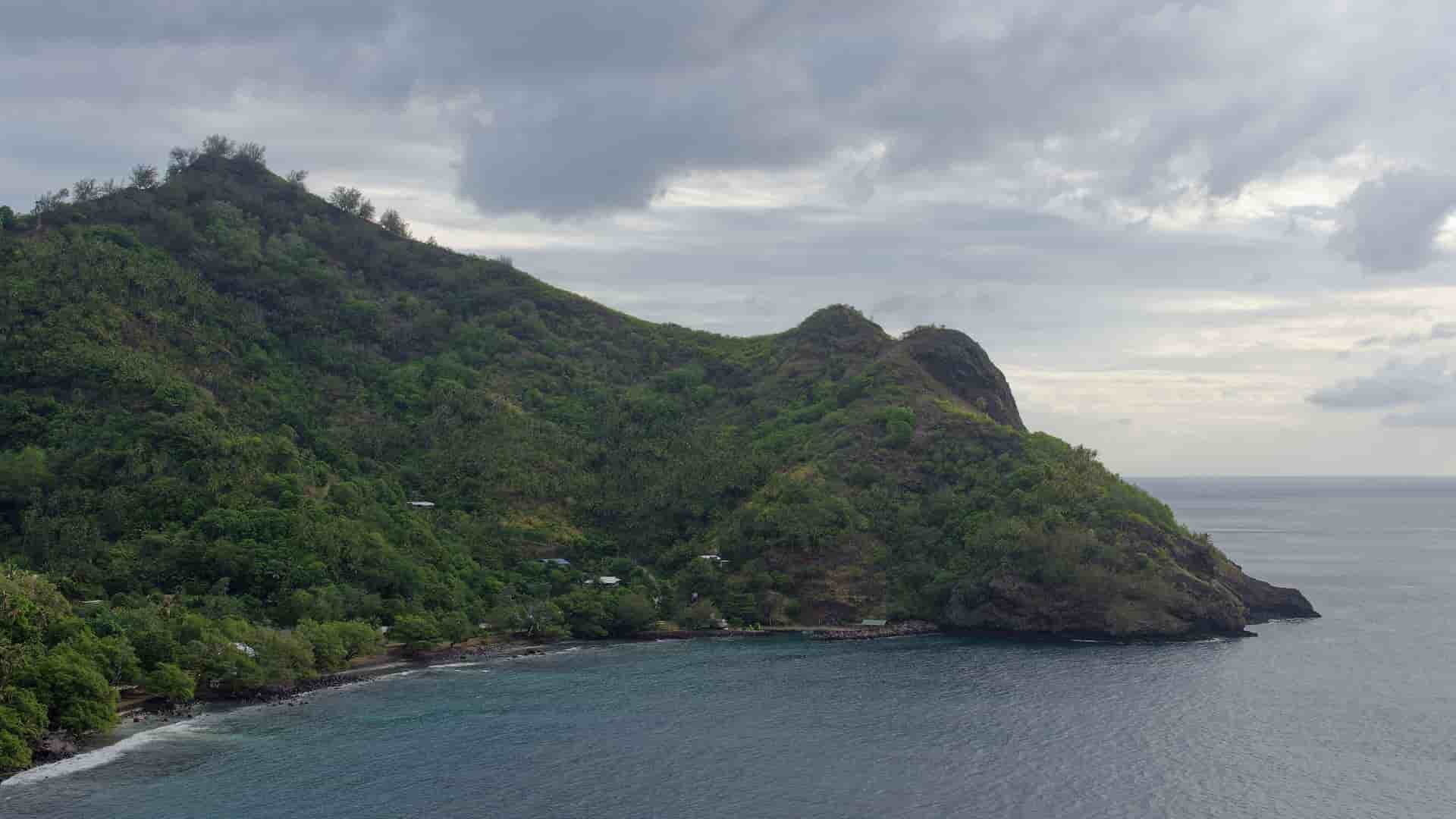 A wide-angle shot of the Hapatoni coast in Tahuata, Marquesas, with a lush, green mountain sloping down to a rocky beach and the serene blue Pacific Ocean.