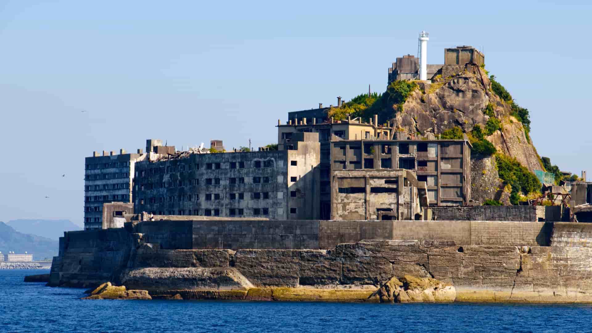 A haunting view of Hashima Island, also known as Gunkanjima, a deserted city with derelict concrete buildings and a lighthouse standing on a rocky outcrop in the sea.