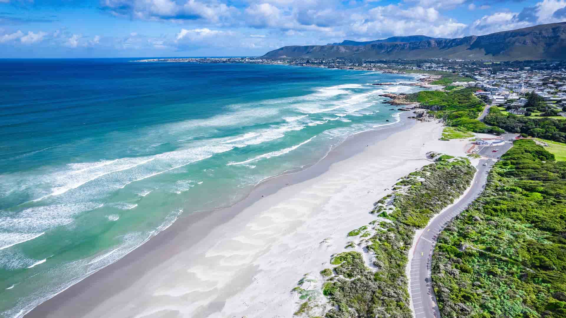"An aerial view of the scenic coastline of Hermanus, South Africa, with a long, white sand beach, turquoise ocean waves, and a winding road along the lush, green shore. "