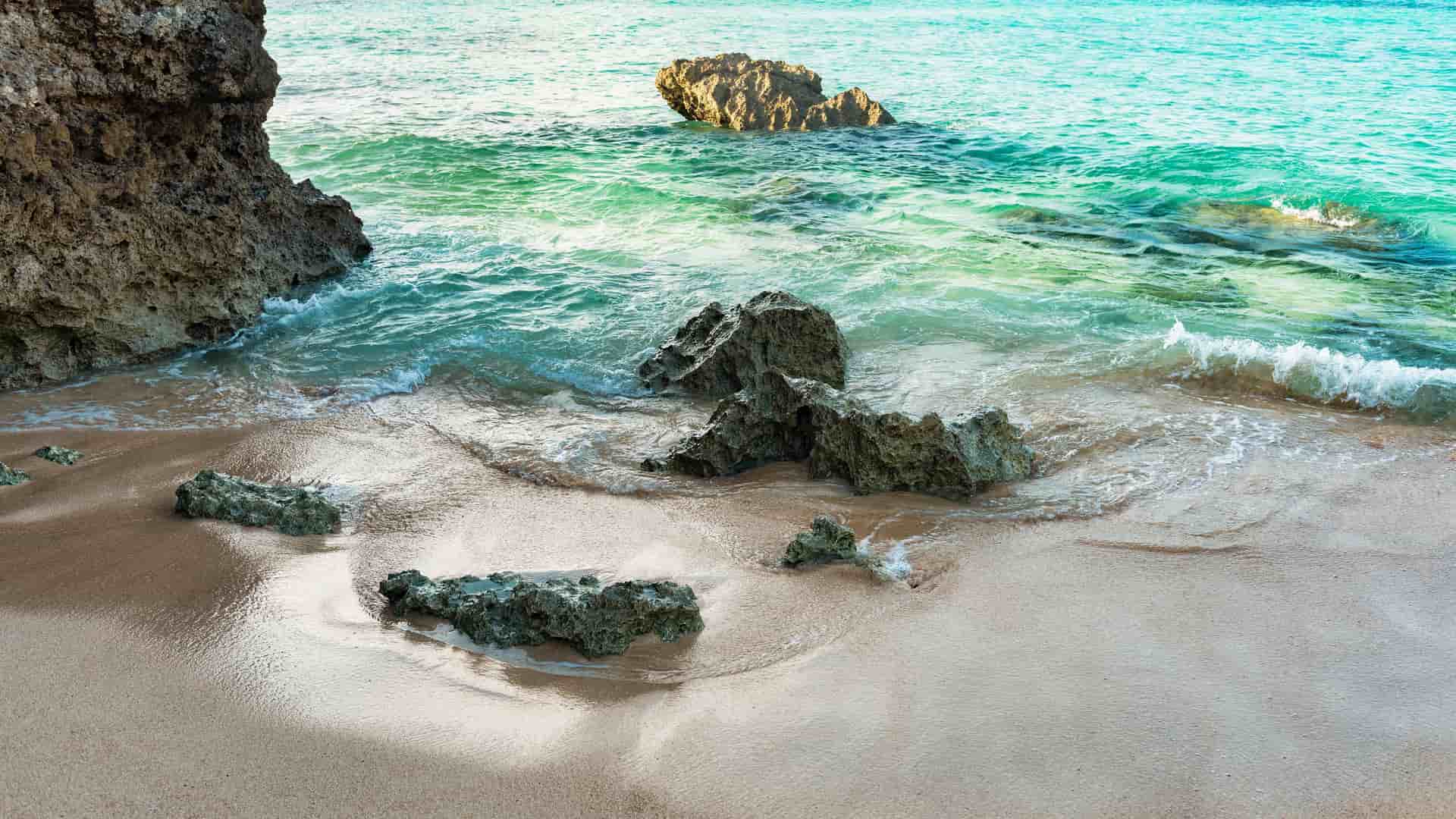 A tranquil view of a sandy beach in Hirara, Miyakojima, with gentle waves from the turquoise water lapping at the shore, and several large rocks scattered on the sand and in the water.