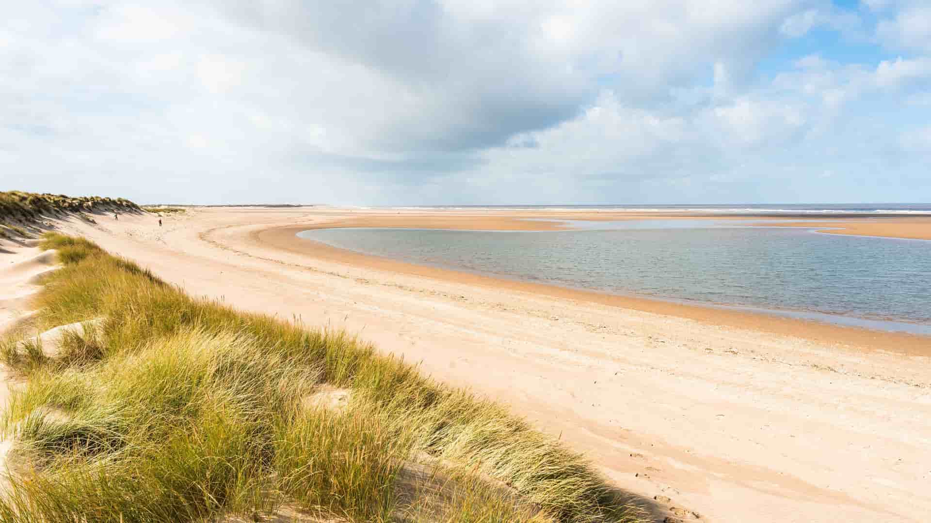 A panoramic view of the vast, sandy expanse of Holkham Beach in Norfolk, England. The image shows a serene tidal lagoon, rolling dunes covered with marram grass, and a wide blue sky with scattered clouds.