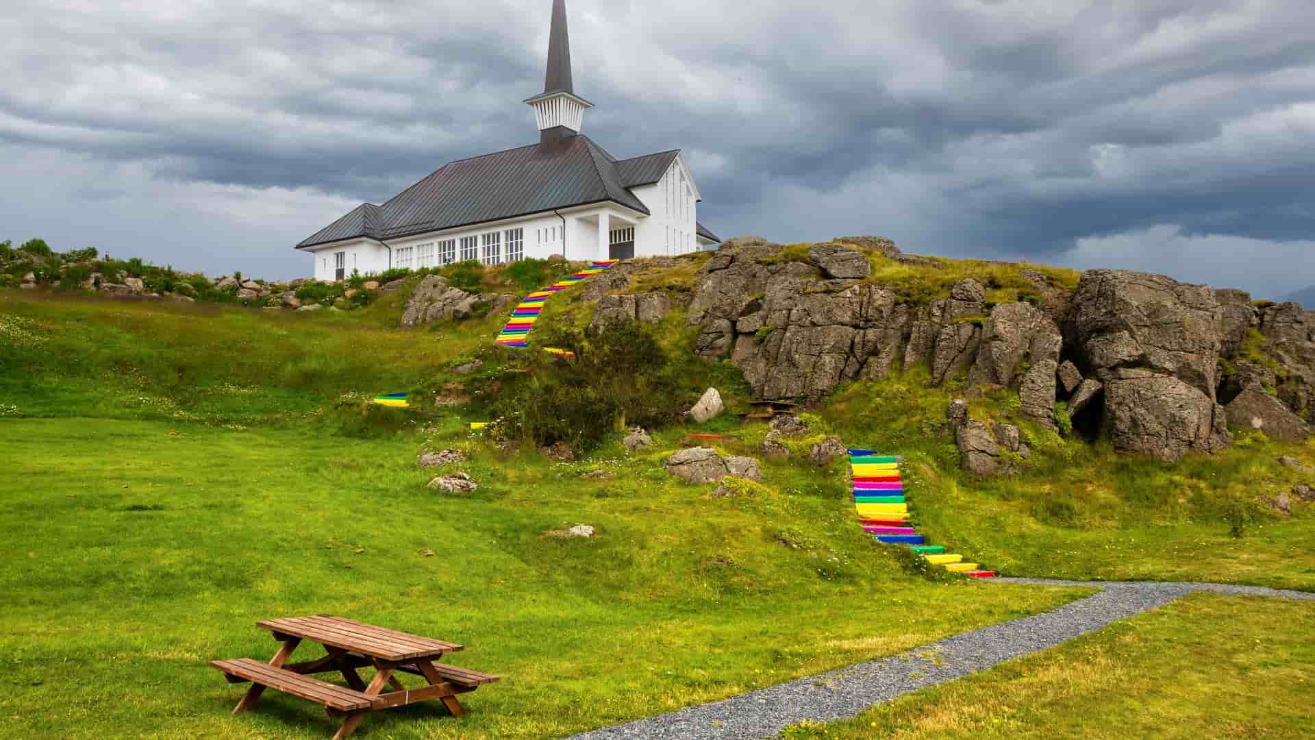 A unique, wide-angle shot of the white wooden church of Holmavik, Iceland, sitting atop a rocky hill. The image is notable for the vibrant rainbow-colored stairs leading up to the church, set against a dramatic, cloudy sky.