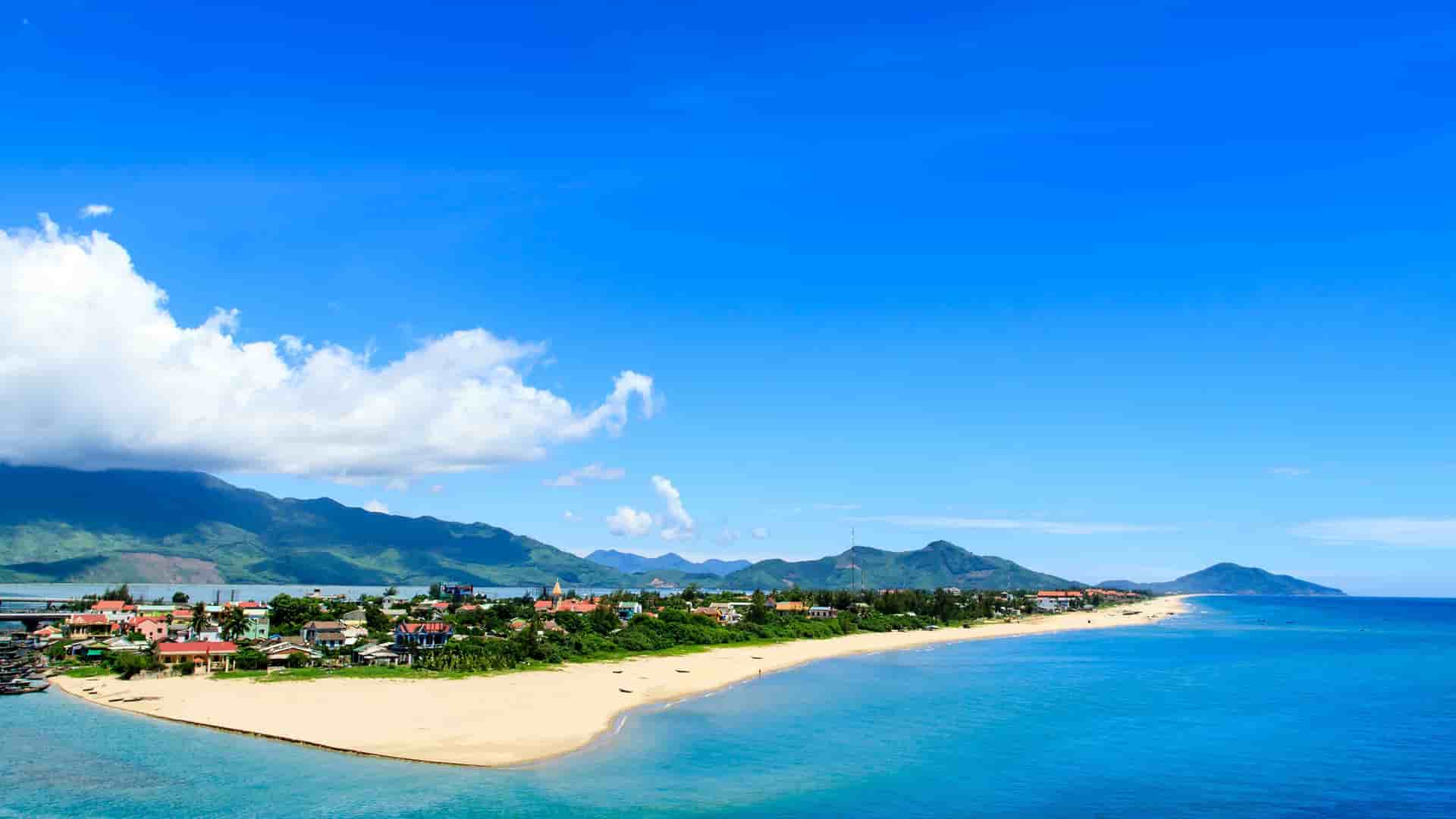 A panoramic shot of a beautiful beach and coastline in Hue, Vietnam, with a small town and mountains in the background, all under a vast, bright blue sky.
