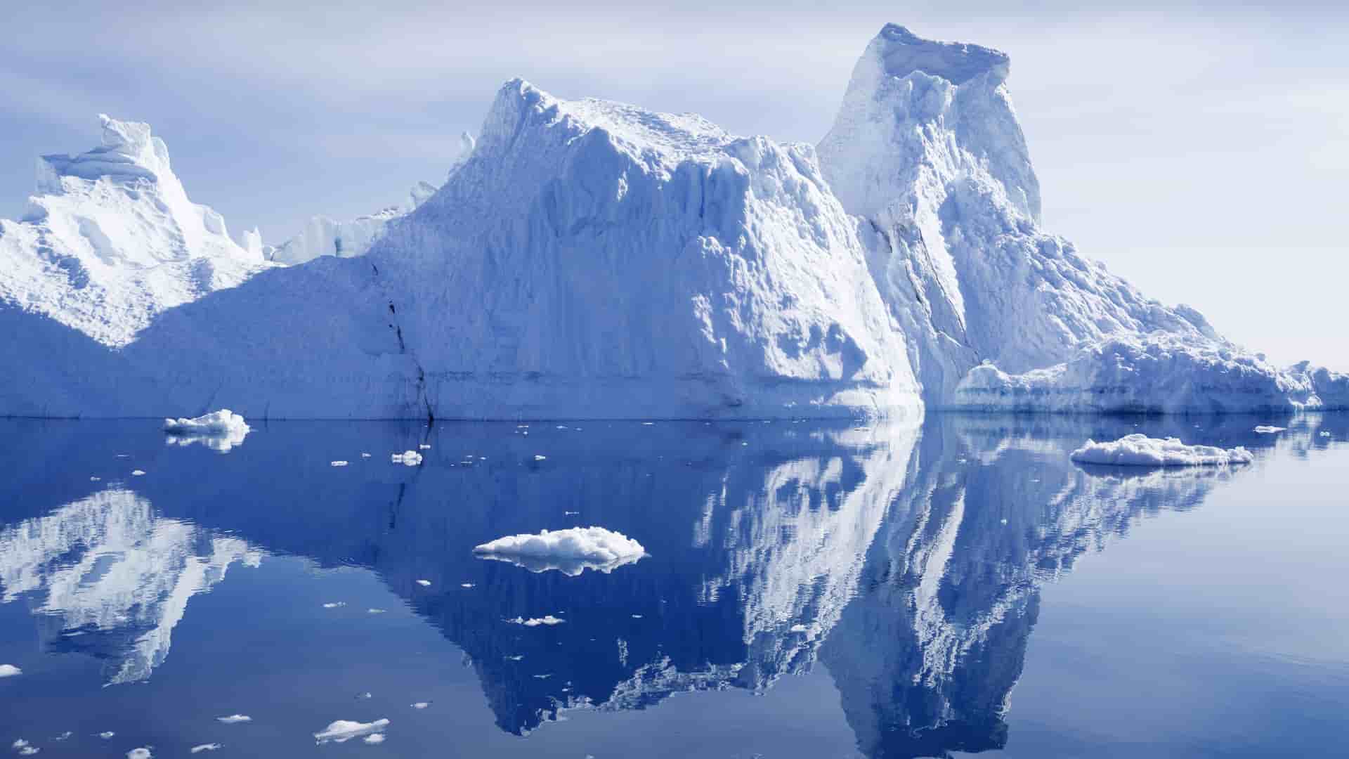 A massive white iceberg with jagged peaks and cliffs, floating in a calm, dark blue sea near Hvalsey, Greenland, with a perfect reflection on the water's surface.