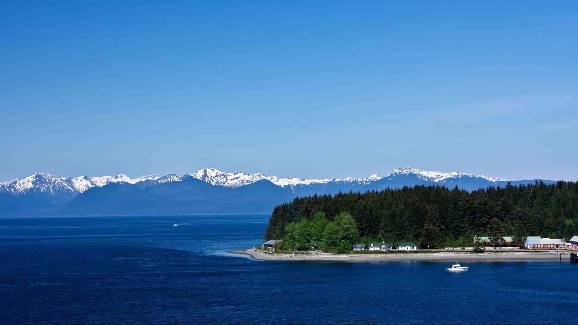 A serene coastal landscape of Icy Strait Point, Alaska, with a small settlement nestled among a dense pine forest, a calm blue sea, and a stunning backdrop of snow-capped mountains.