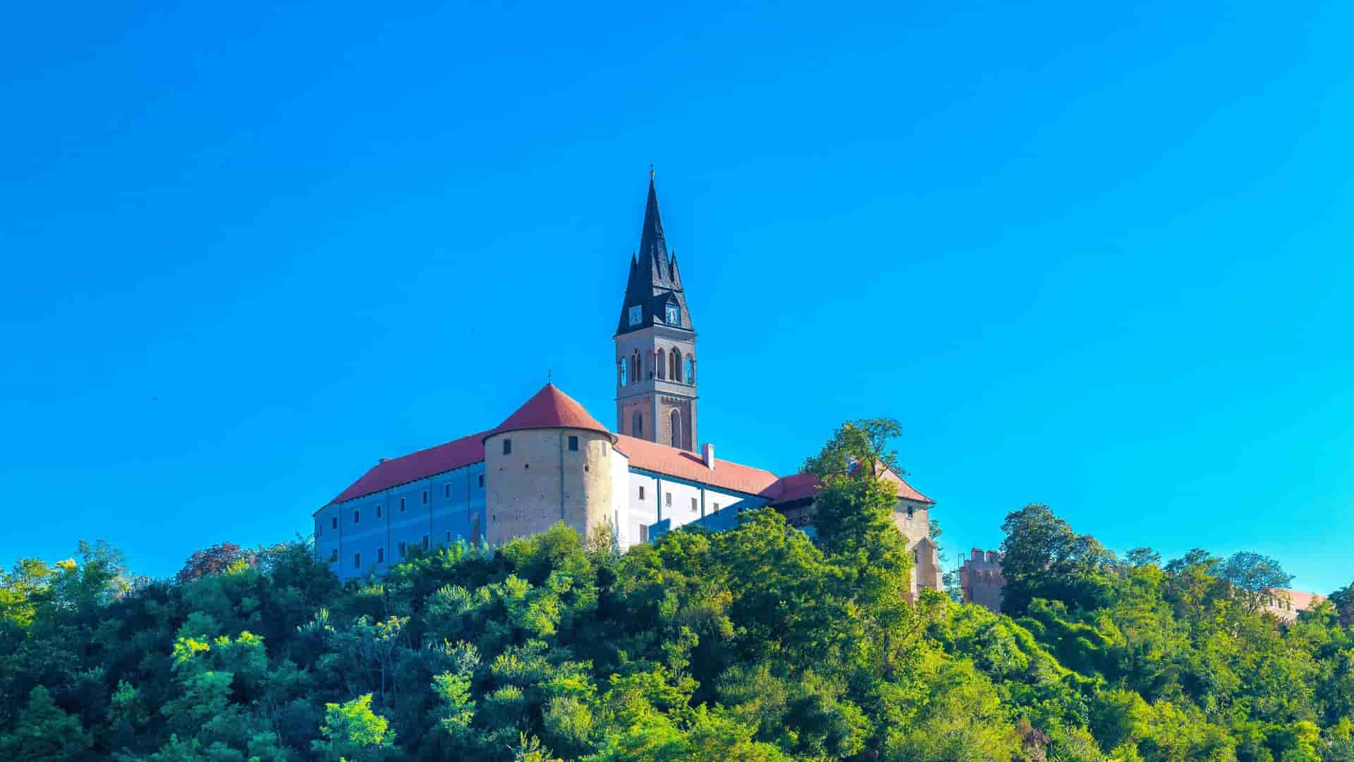 A majestic shot of Ilok Castle in Croatia, showcasing the large fortress with its prominent bell tower and red-roofed buildings perched on a lush, green hill under a clear blue sky.