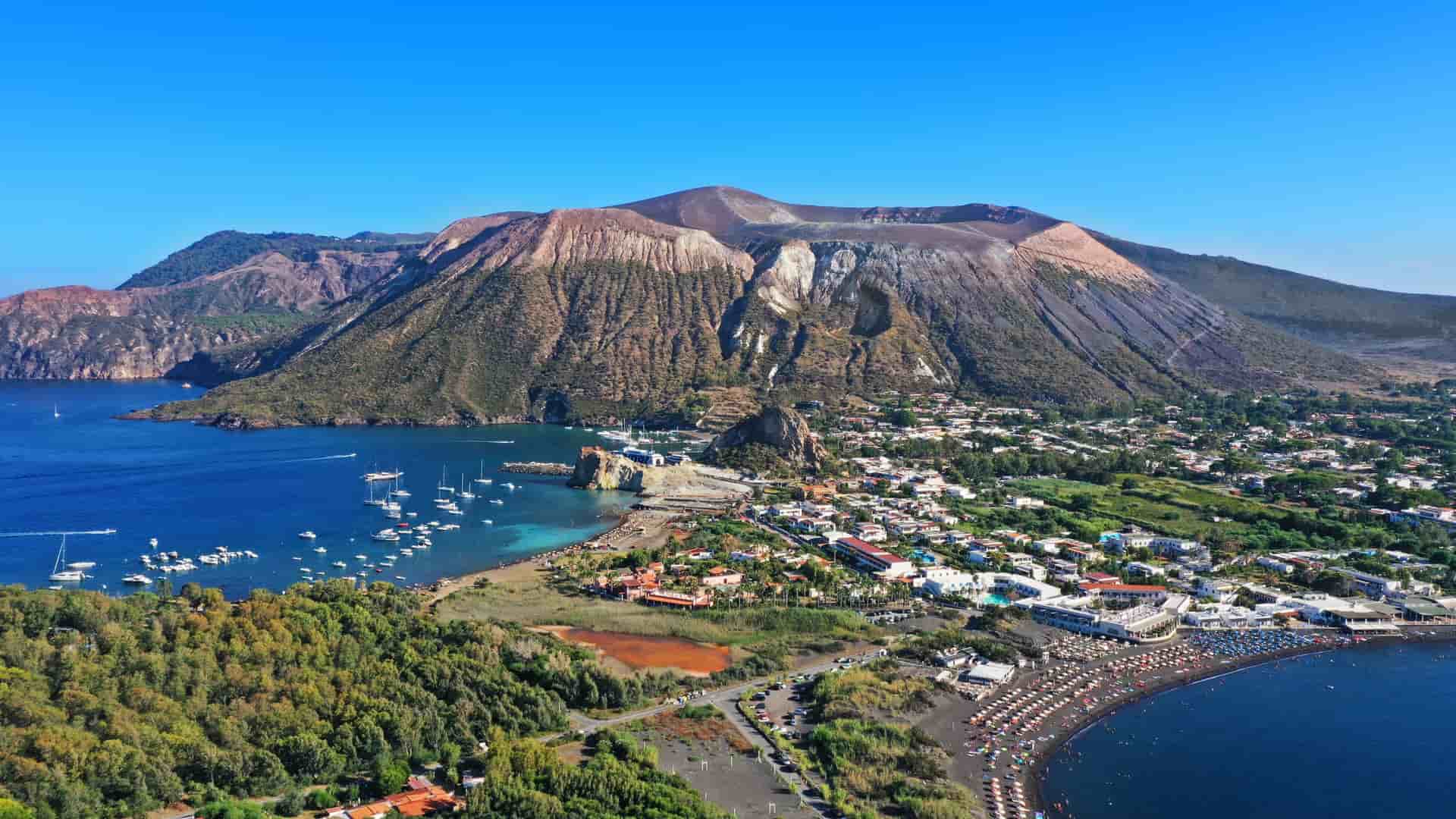 An aerial view of Isola di Stromboli, an active volcano with its summit visible, overlooking a town with a black sand beach and boats in the bay.