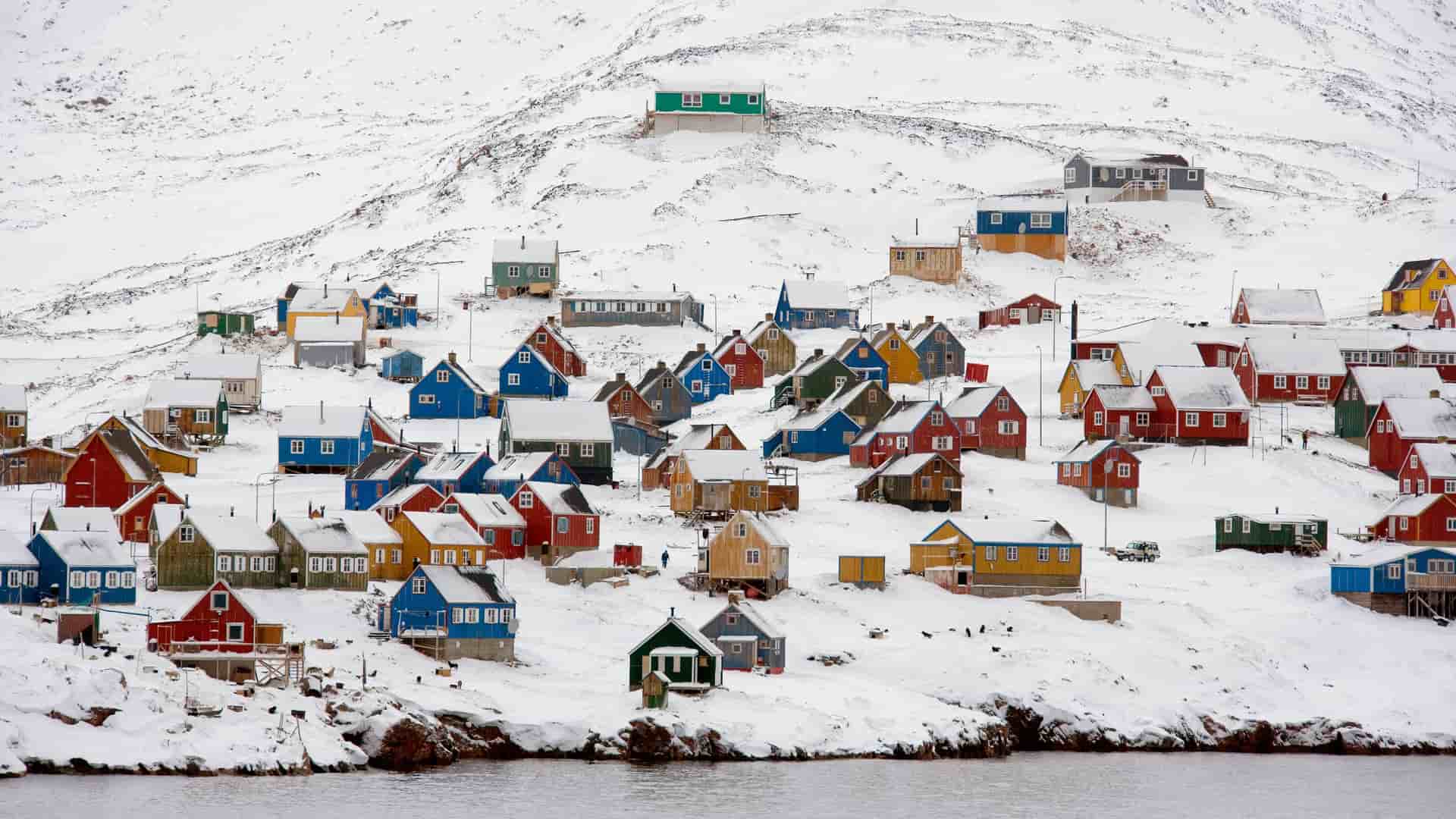 A winter wonderland view of the remote Ittoqqortoormiit village in Greenland, with colorful houses nestled on a snow-covered hillside overlooking the water.