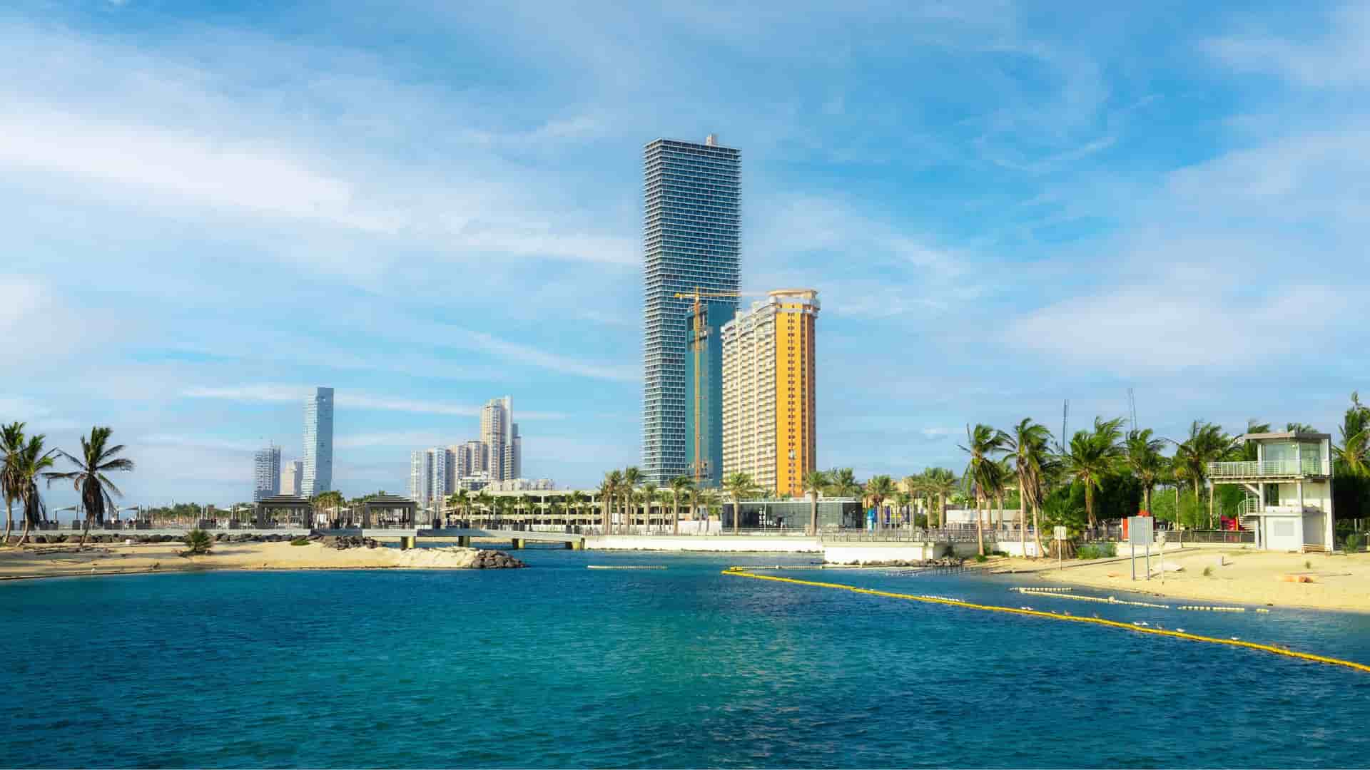 A scenic view of Jeddah's modern skyline, with towering skyscrapers and lush palm trees lining a sandy beach and a clear blue bay.