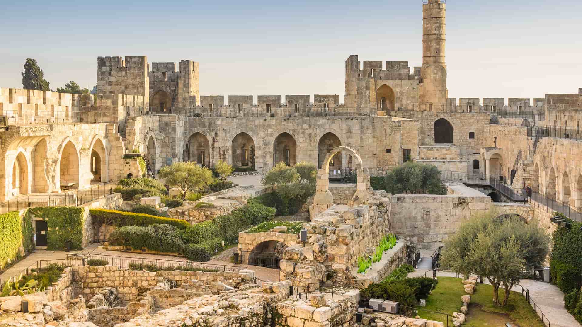 "A historic shot of the Citadel of David (Tower of David) in Jerusalem, Israel, showing the ancient stone walls, arches, and a minaret, with green gardens and archaeological ruins in the foreground.  "