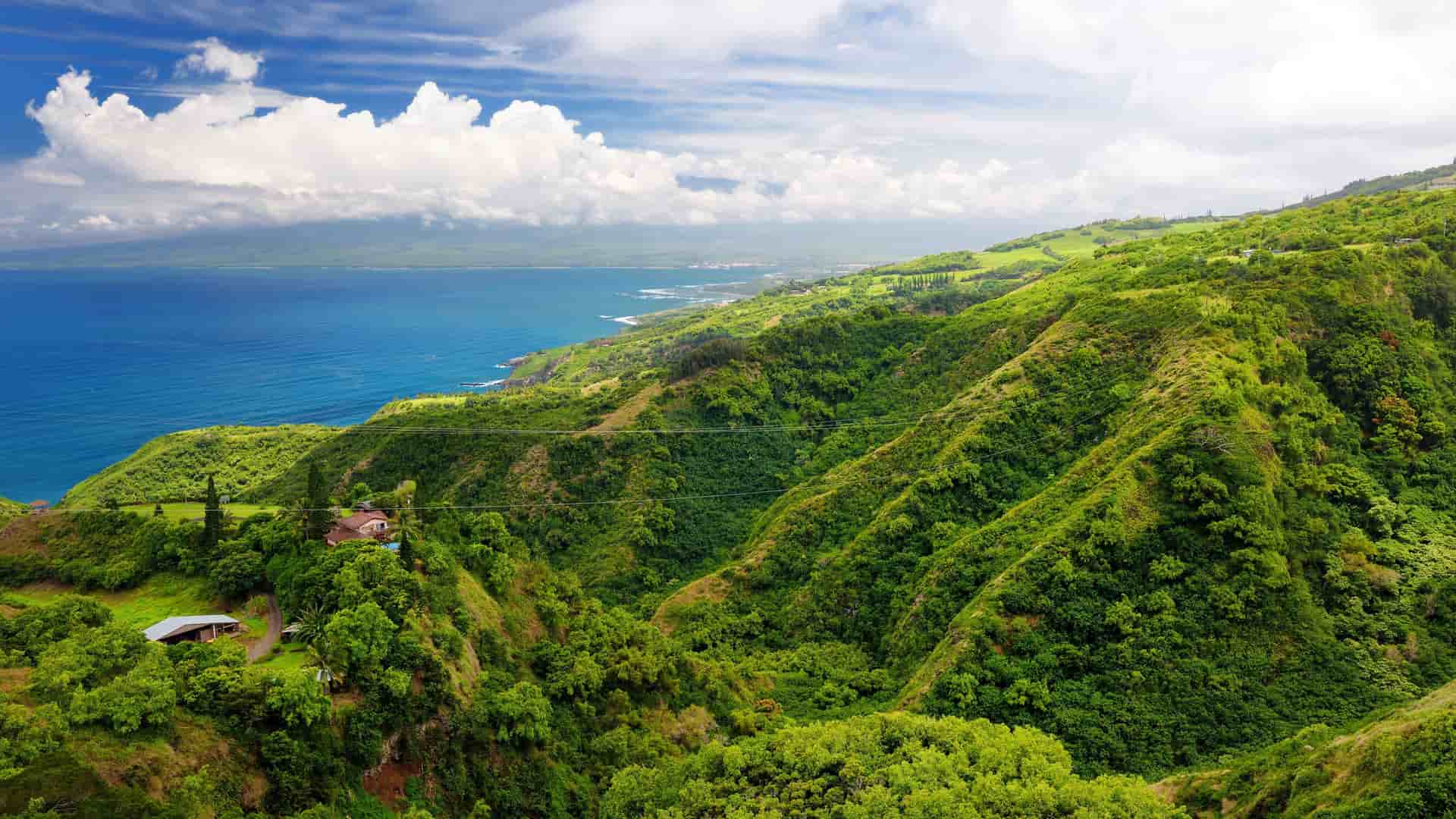 A stunning panoramic view of the lush, green hills and valleys of Maui, with the deep blue Pacific Ocean and coastline stretching into the distance under a partly cloudy sky near Kahului, Hawaii.