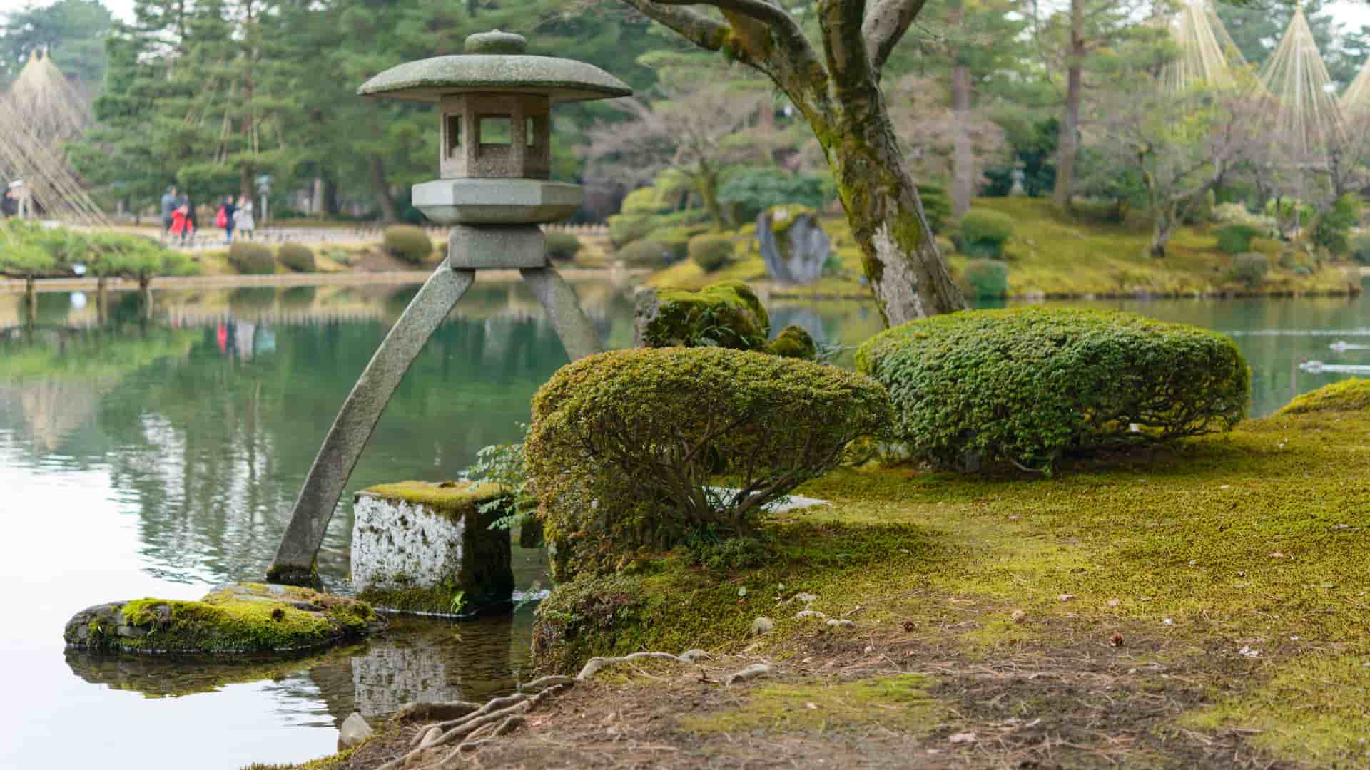 A beautiful view of the historic Kenrokuen Garden in Kanazawa, Japan, featuring the iconic Kotoji-toro lantern on the edge of a pond with lush green moss and manicured bushes.