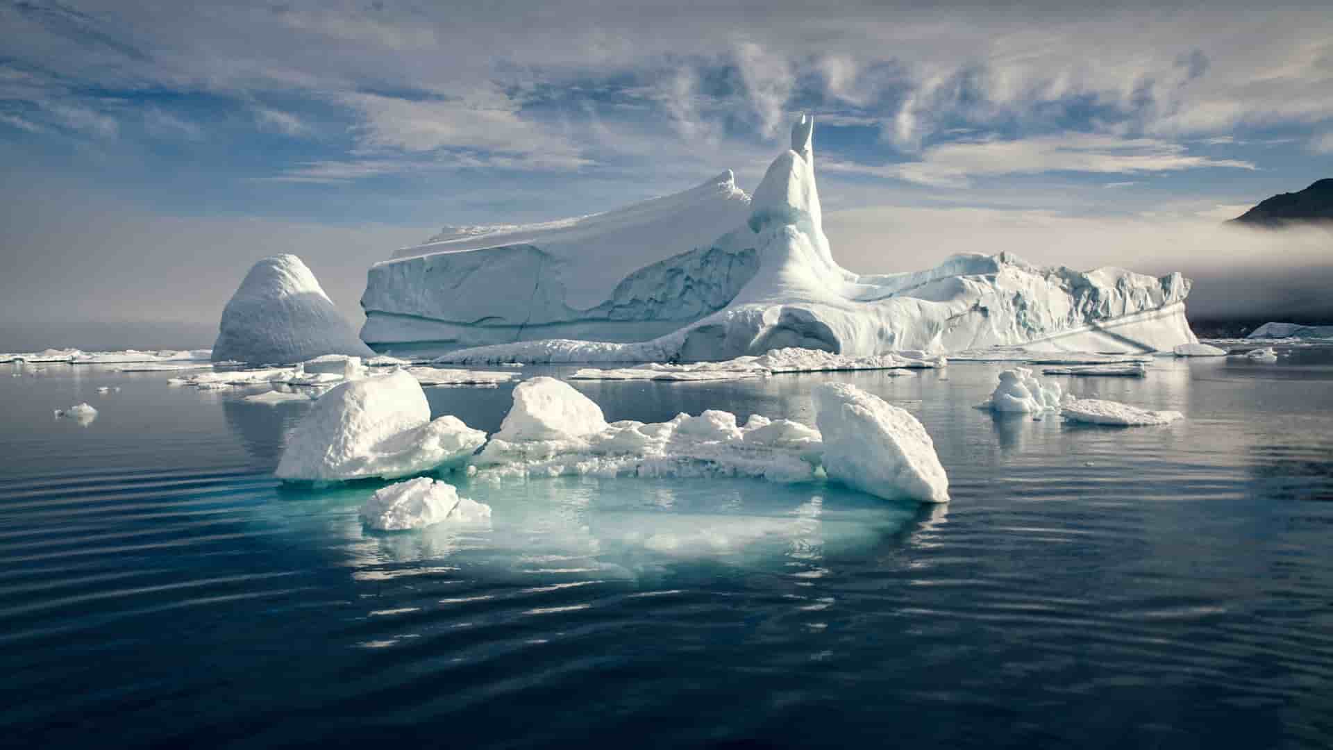 A wide-angle landscape of icebergs floating in the Kangerlussuaq Fjord in Greenland, with one massive, uniquely shaped iceberg dominating the scene under a dramatic sky.