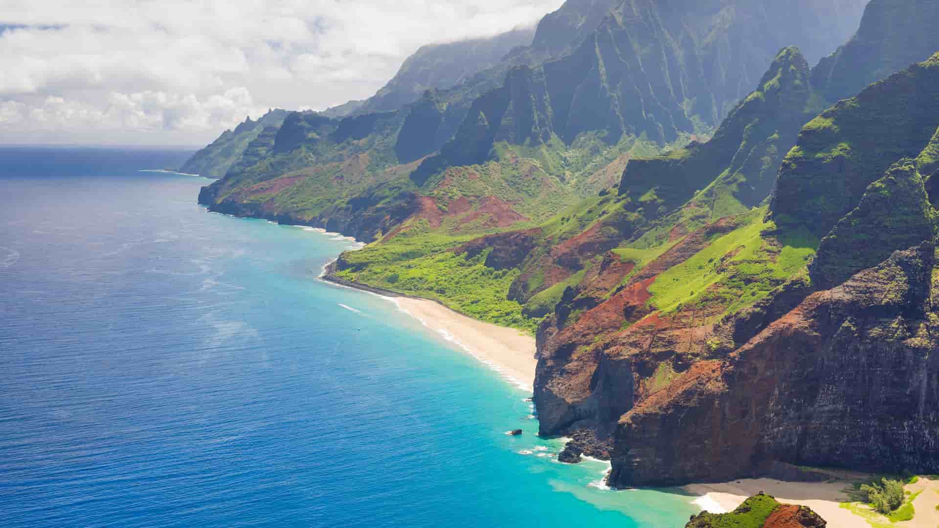 An aerial view of Kauai's rugged Na Pali Coast, showcasing lush green cliffs, a pristine secluded beach, and the vibrant blue Pacific Ocean.