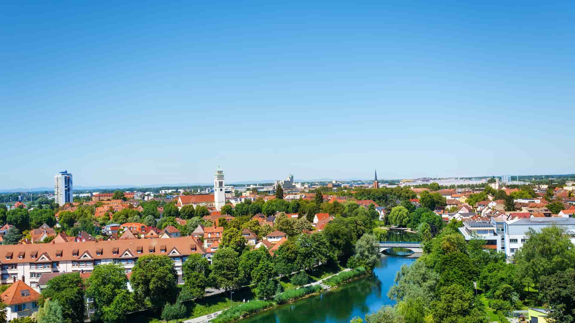 A scenic panoramic view of Kehl, Germany, with a winding river in the foreground, lush green trees, and the city's red-roofed buildings under a clear blue sky.