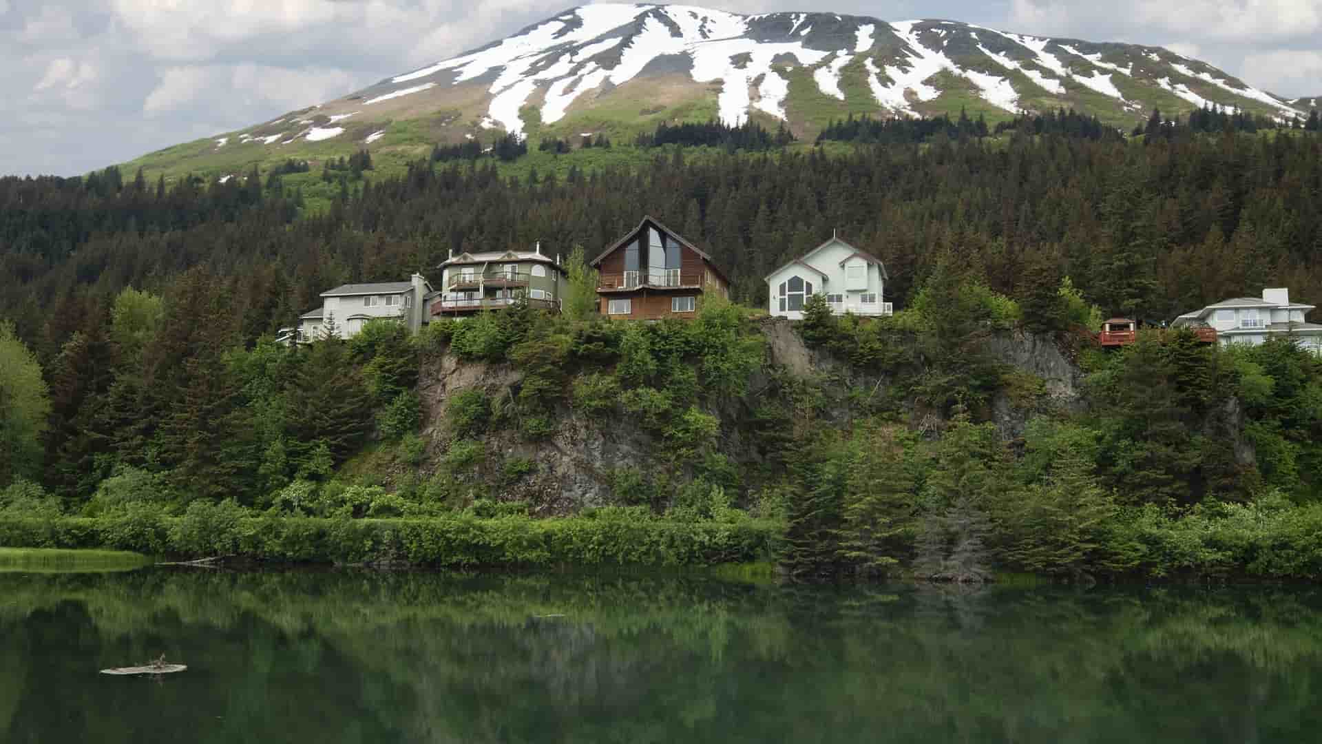 A beautiful waterfront view of the Kenai Princess Wilderness Lodge nestled on a cliffside, surrounded by thick forest with a snow-capped mountain in the background.
